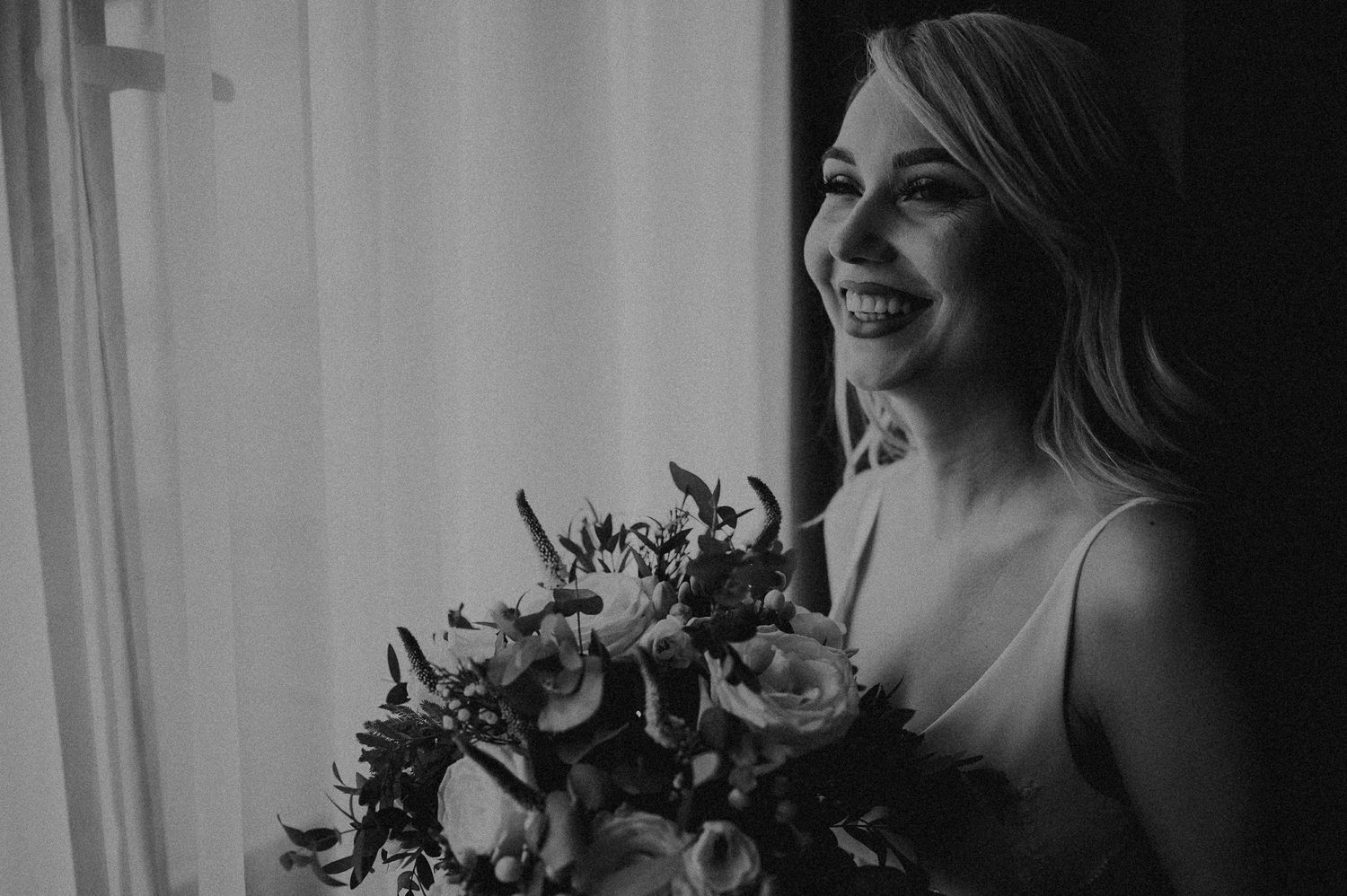 Black and white portrait of smiling bride holding colorful floral bouquet near bright window.