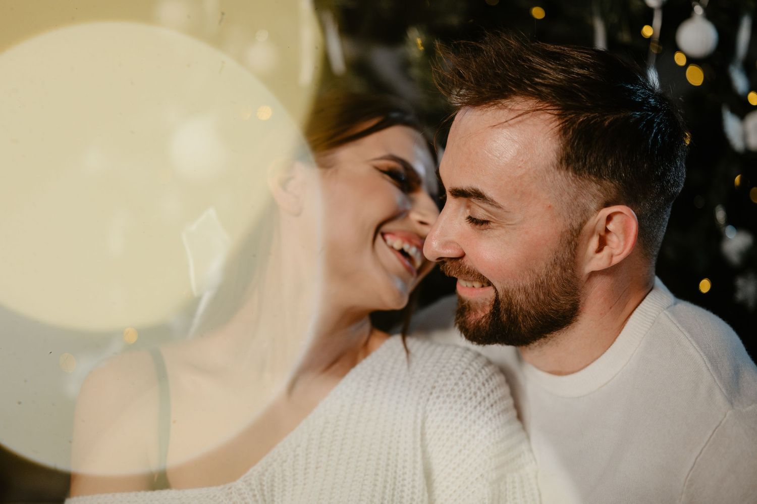 Romantic couple laughing together near sparkling Christmas tree with golden bokeh lights.