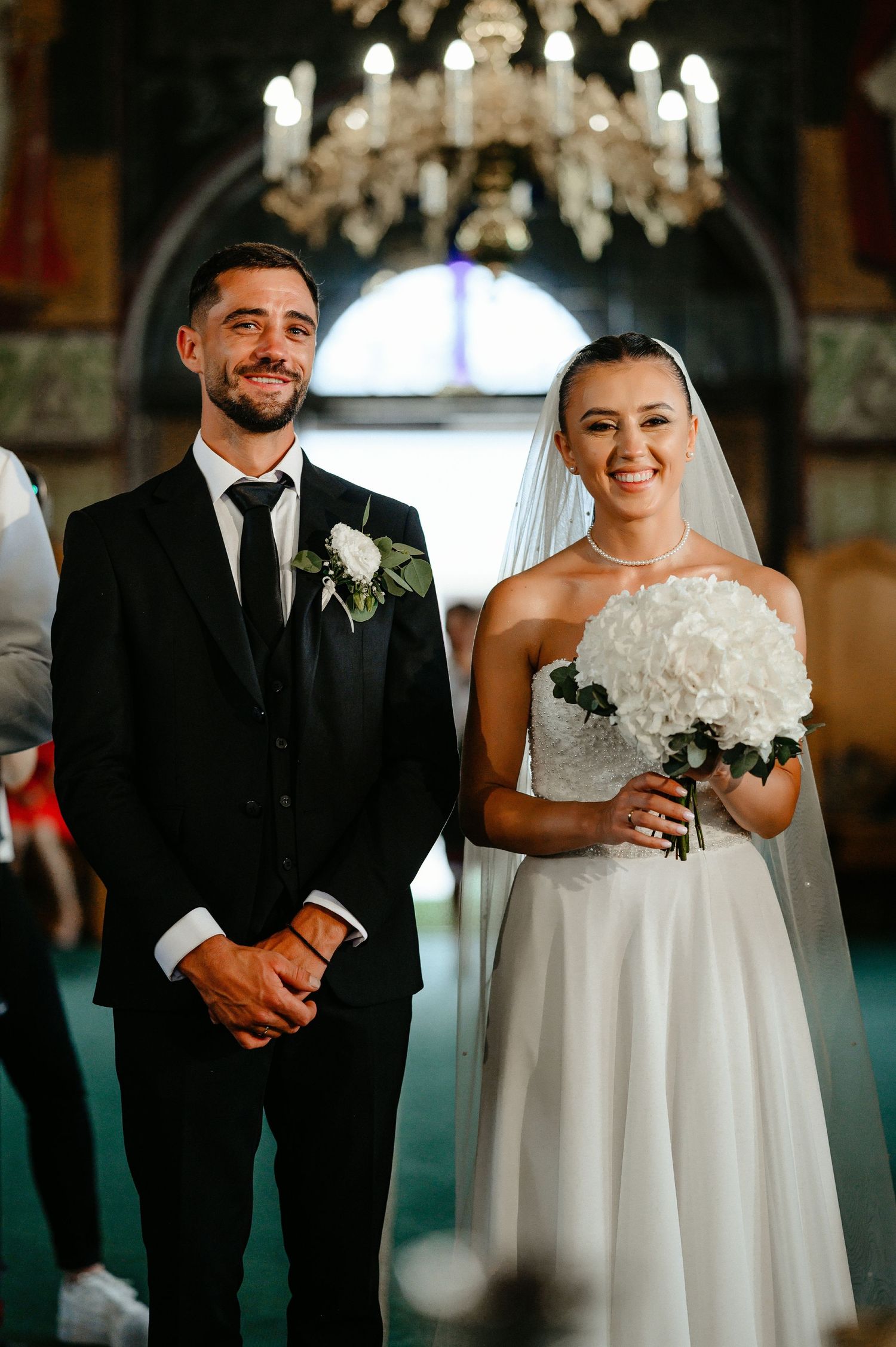Happy bride and groom smile together holding white bouquet during Orthodox church wedding ceremony.