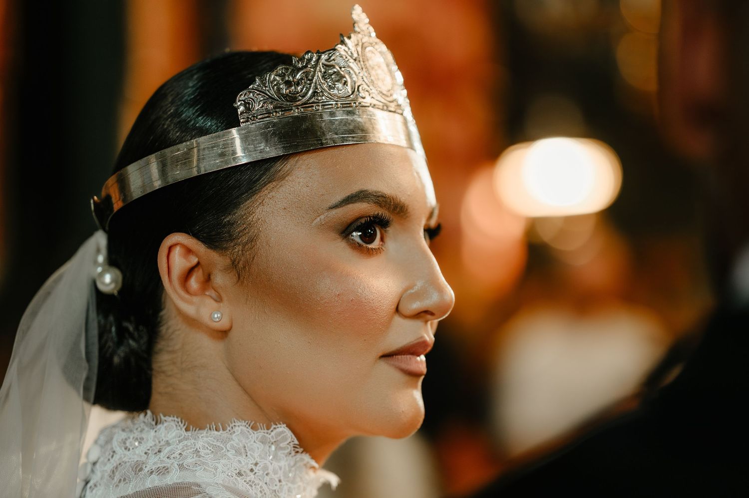 Beautiful bride wearing ornate silver wedding crown and veil during Orthodox church ceremony.