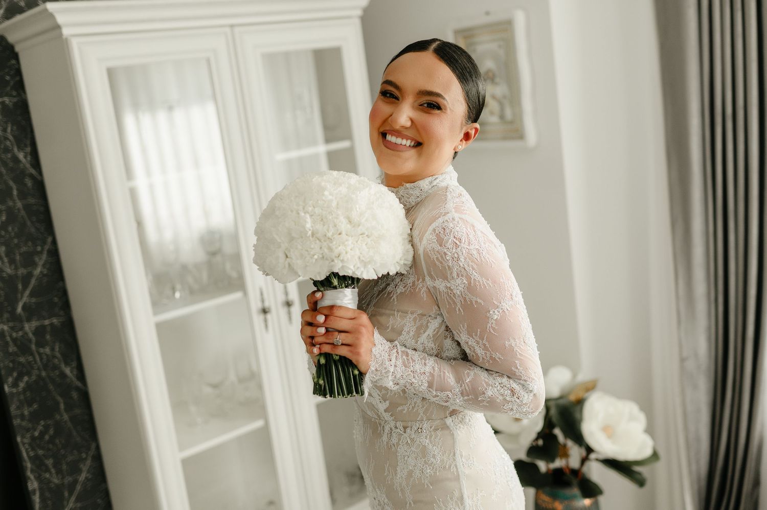 Smiling bride in lace high-neck wedding dress holding white hydrangea bouquet indoors.