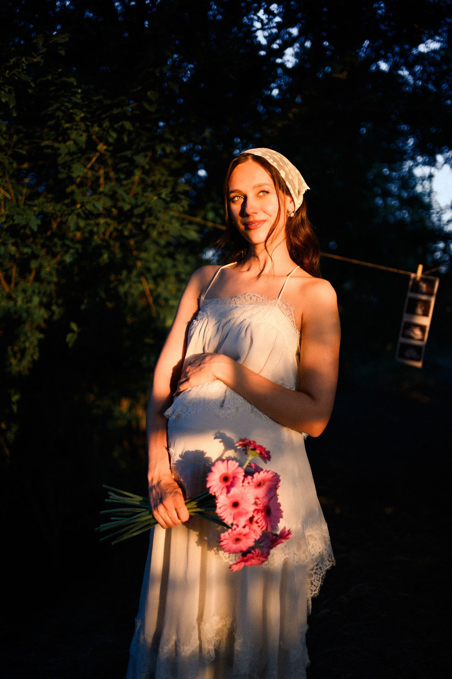 Pregnant woman in white dress holding pink gerbera daisies, wearing lace headband, bathed in golden sunset light outdoors.