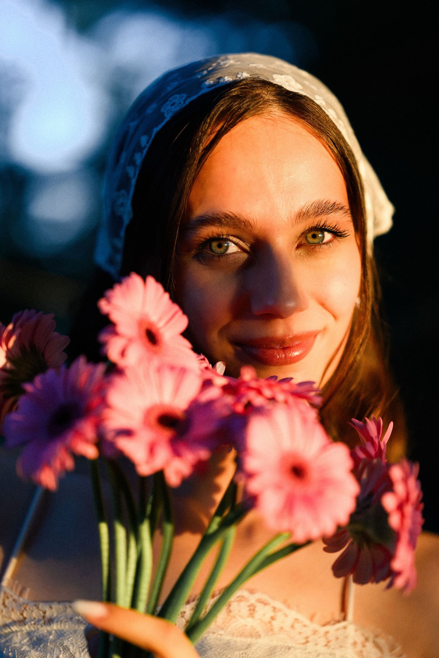 Young woman in white headscarf holding pink gerbera daisies, bathed in warm golden hour light.
