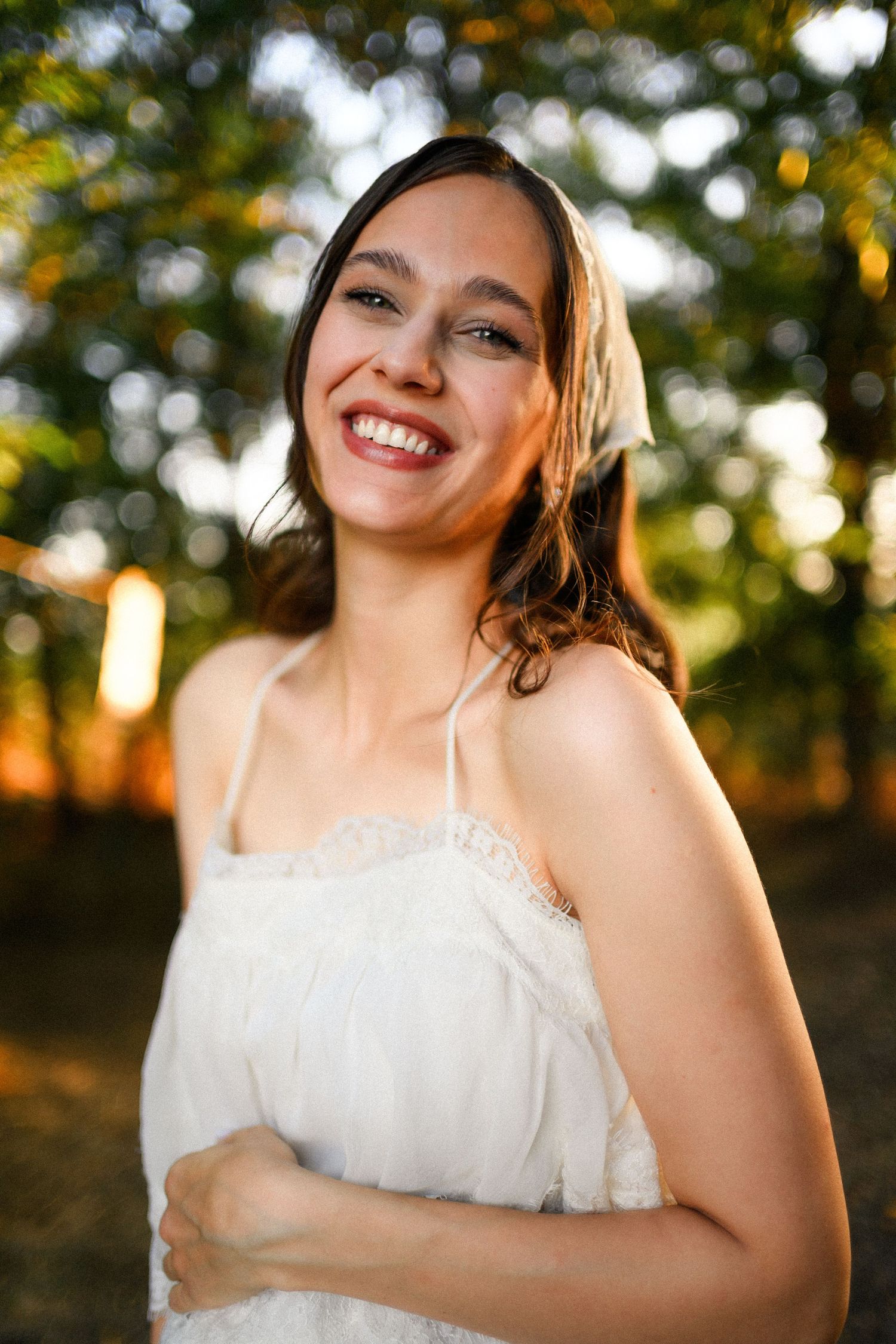 Smiling young woman in white sundress outdoors with golden sunset light filtering through green trees behind her.
