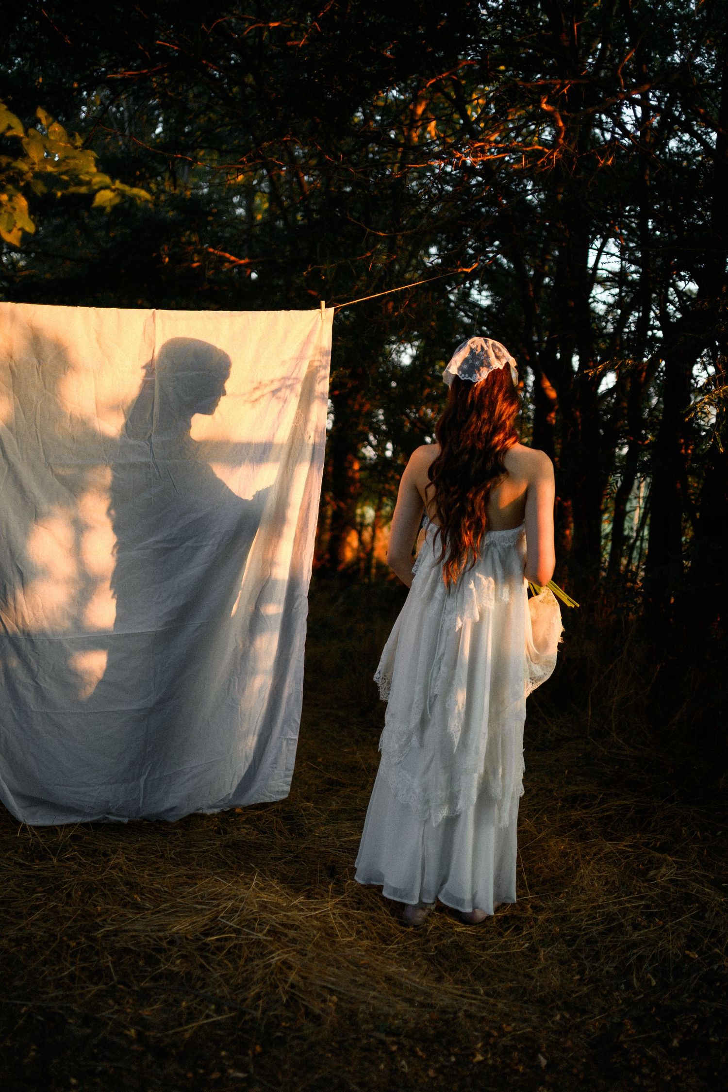 A woman in a white wedding dress stands before a hanging sheet displaying a mysterious shadow silhouette in a forest.