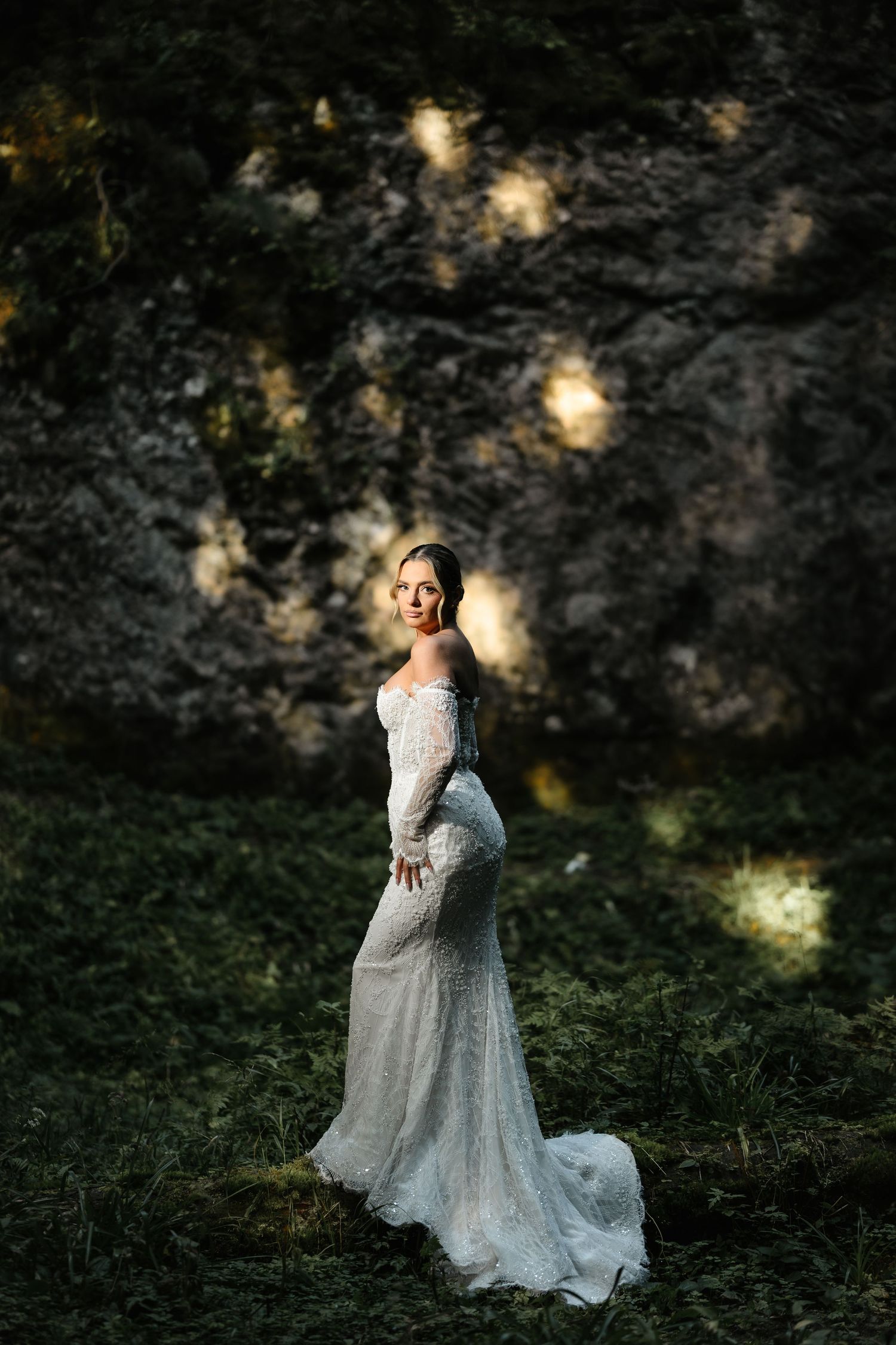 Bride in white lace off-shoulder gown stands in a lush forest setting with dramatic rocky backdrop.