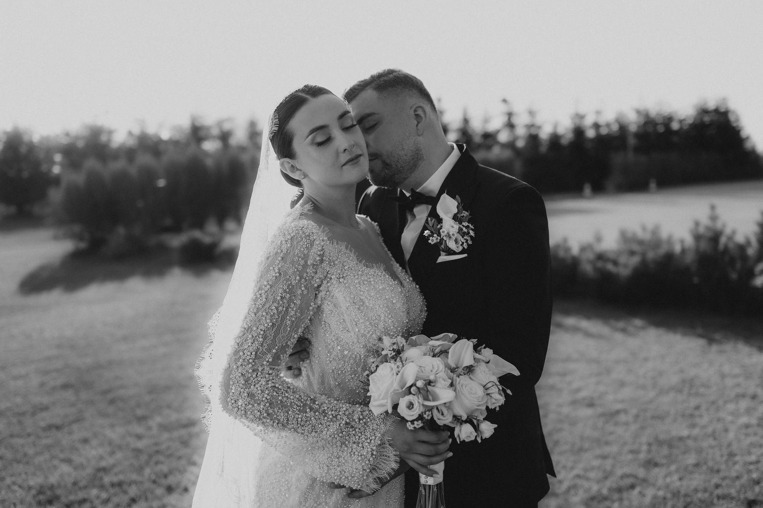 Black and white wedding photo of bride and groom sharing a romantic moment outdoors, bride holding a bouquet.