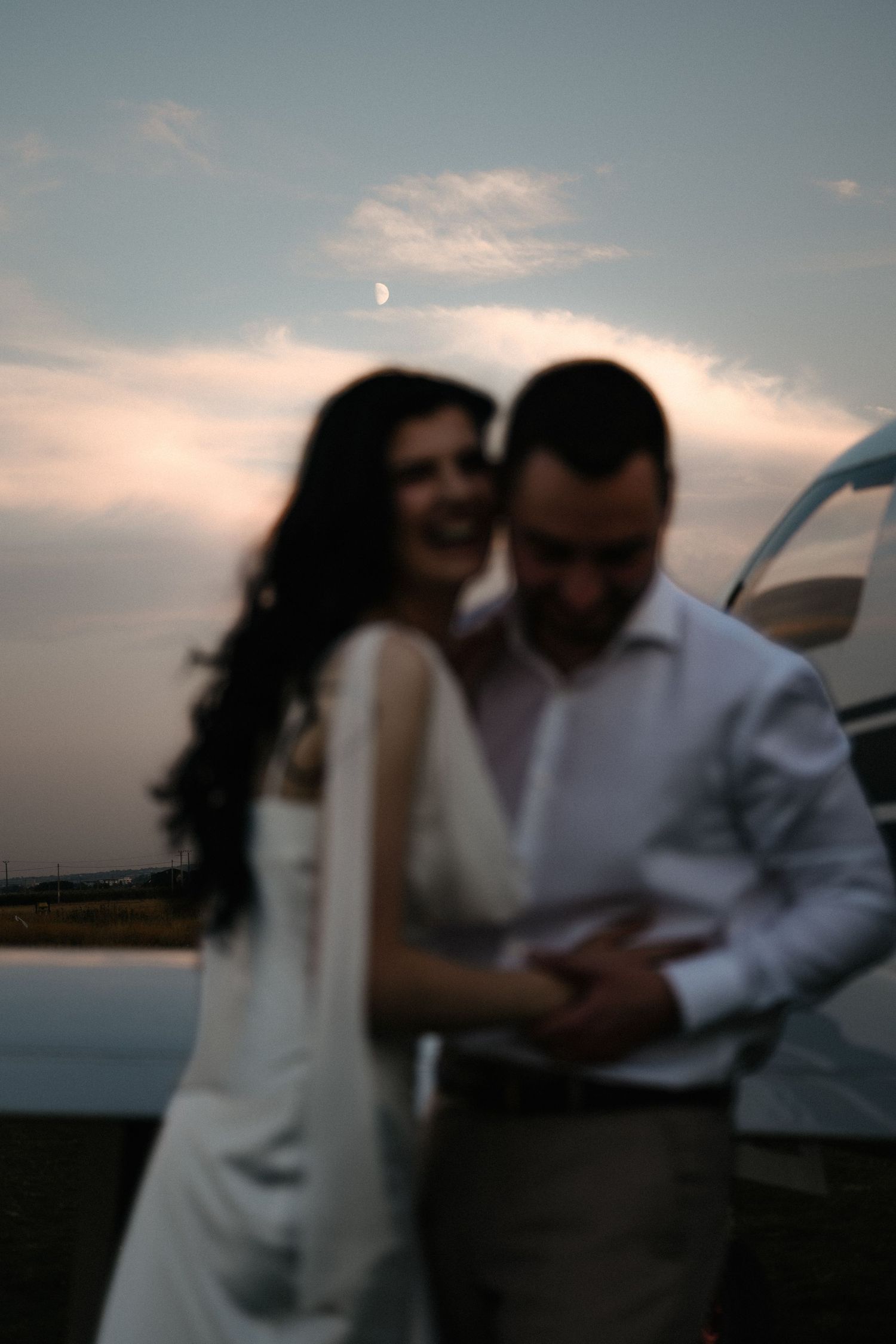 Blurry romantic couple embracing at sunset with crescent moon visible in cloudy sky, woman in white dress smiling.