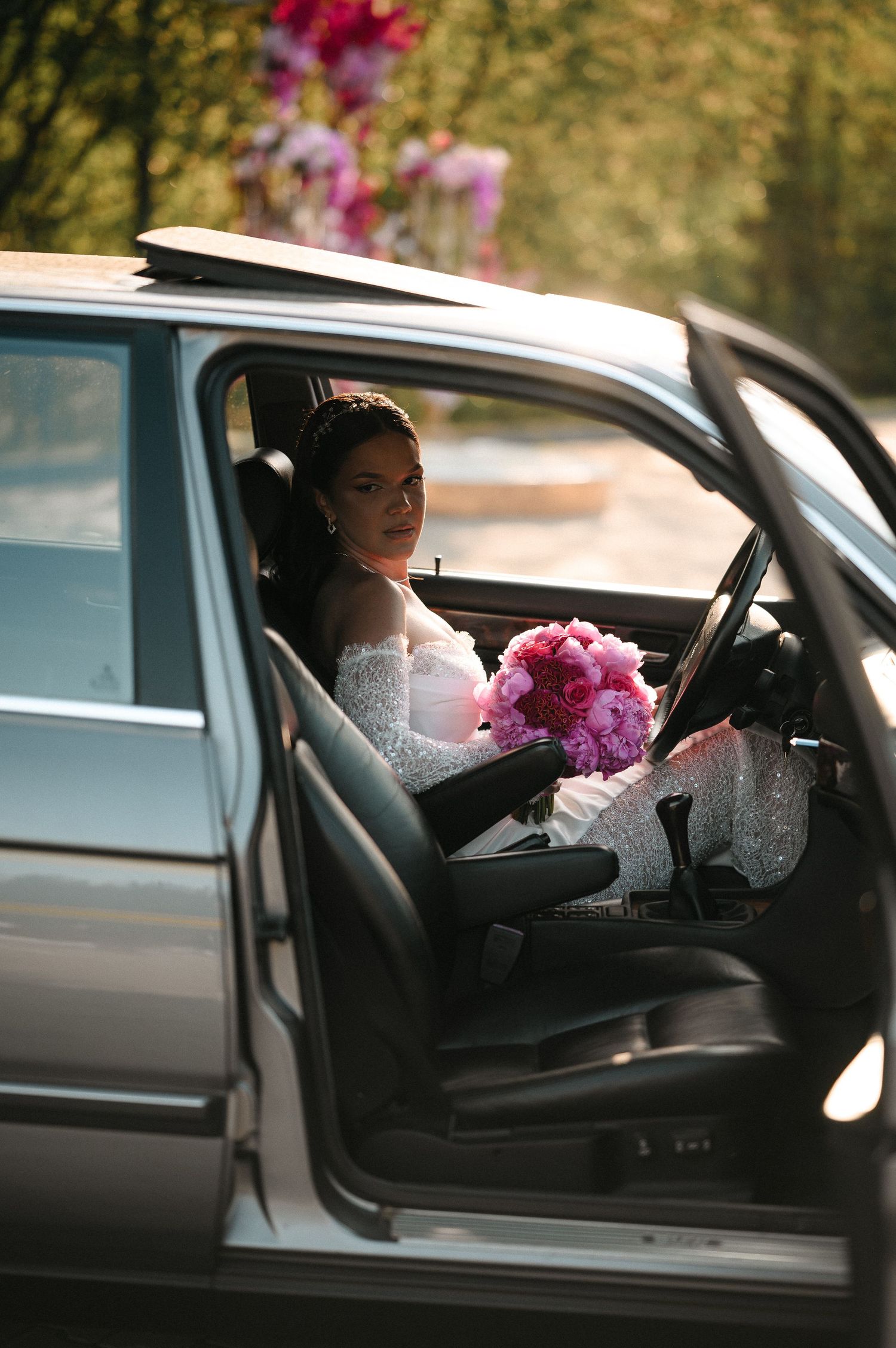 Bride in white gown holding pink peony bouquet seated in luxury car with open door on wedding day.
