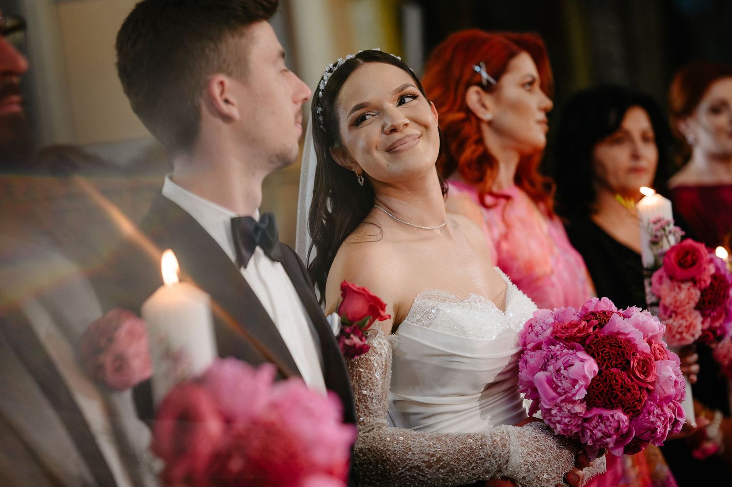 Smiling bride in white gown holding pink peonies beside her groom during a candlelit church ceremony.