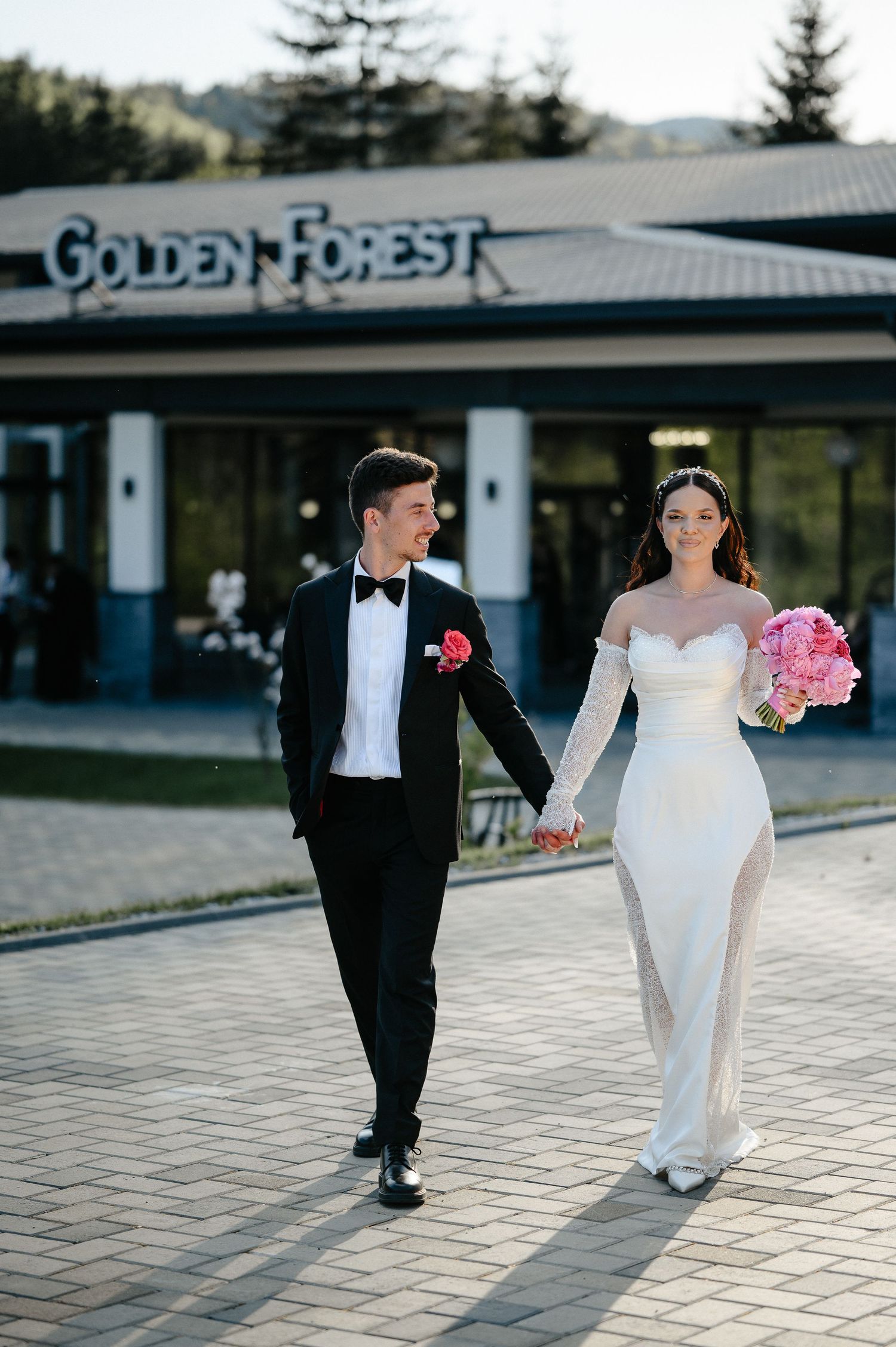 Smiling newlyweds strolling hand in hand outside Golden Forest venue, bride carrying vibrant pink peony bouquet.