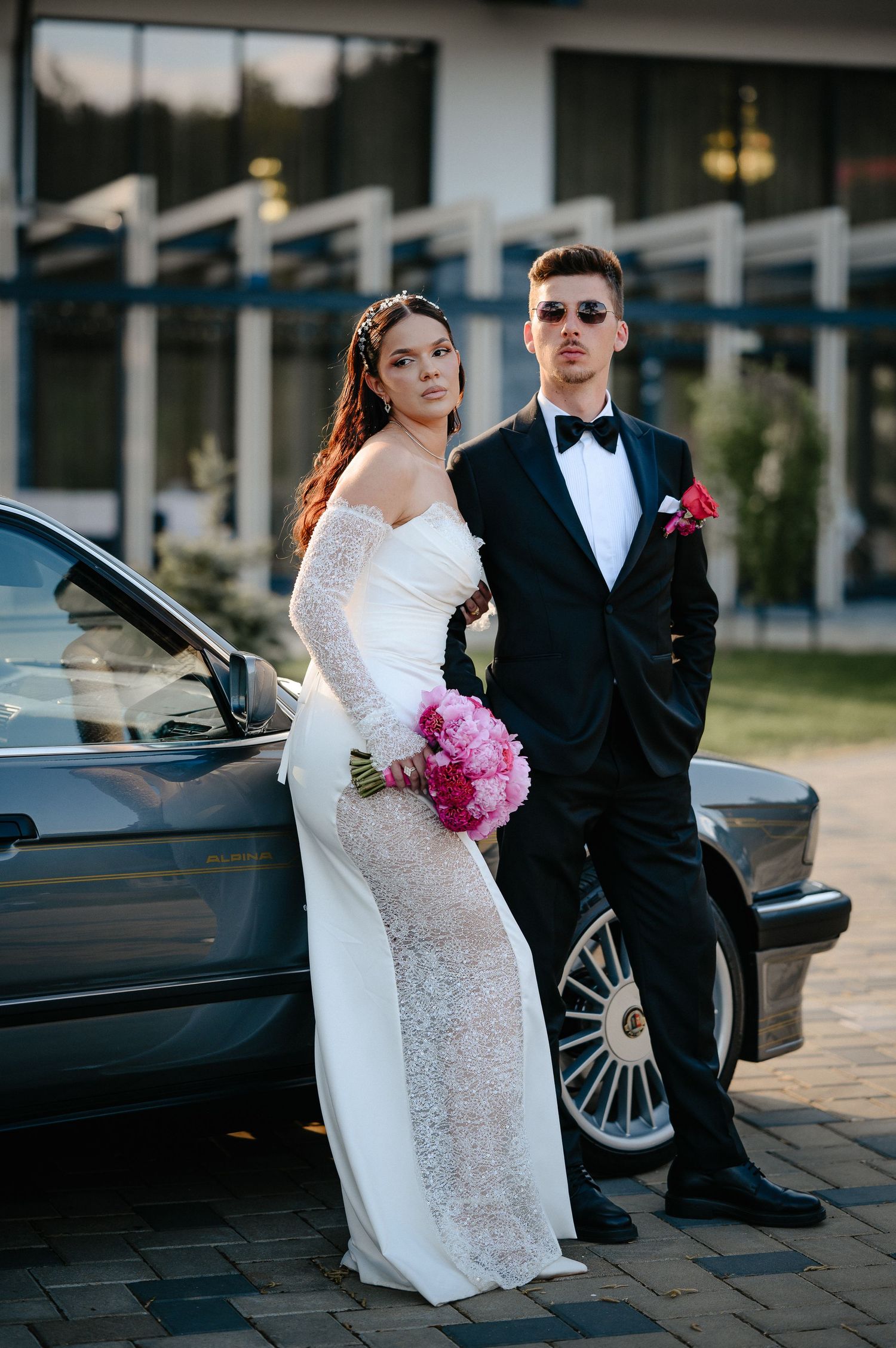 Elegant bride in lace gown holding pink bouquet poses with groom in tuxedo beside classic gray BMW.