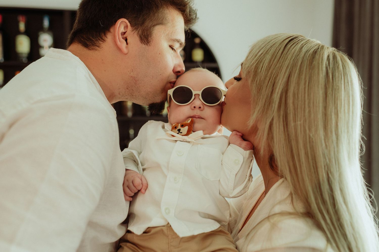Parents kissing their newborn baby wearing cute round sunglasses from both sides simultaneously.