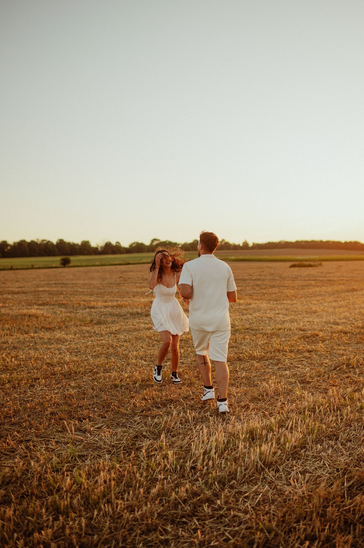 Young couple holding hands running through harvested field during golden hour.
