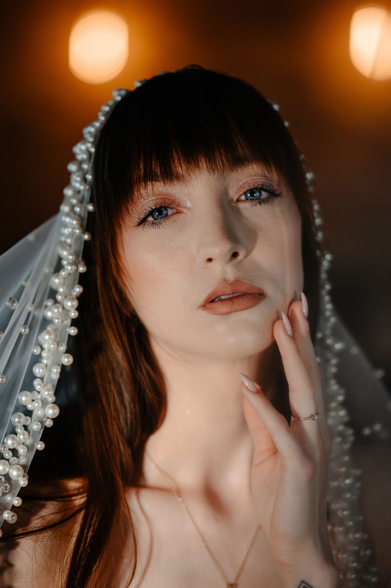 Stunning bride with bangs and pearl-edged veil poses thoughtfully in warm glowing candlelight.