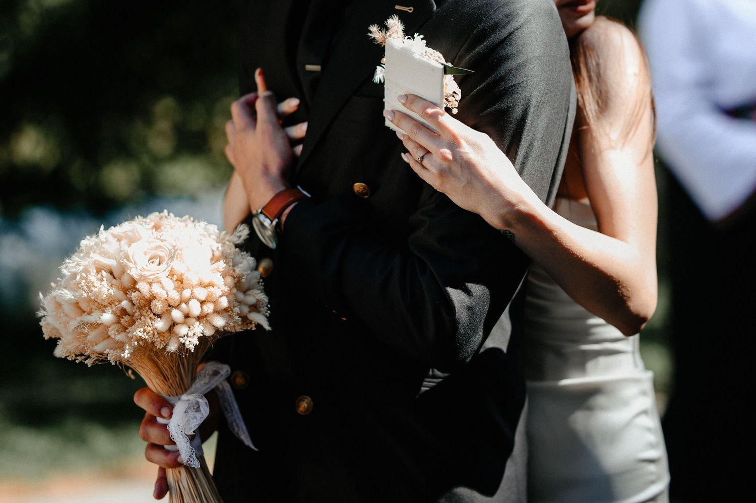 Close-up of groom in black suit holding dried flower bouquet embracing bride outdoors.
