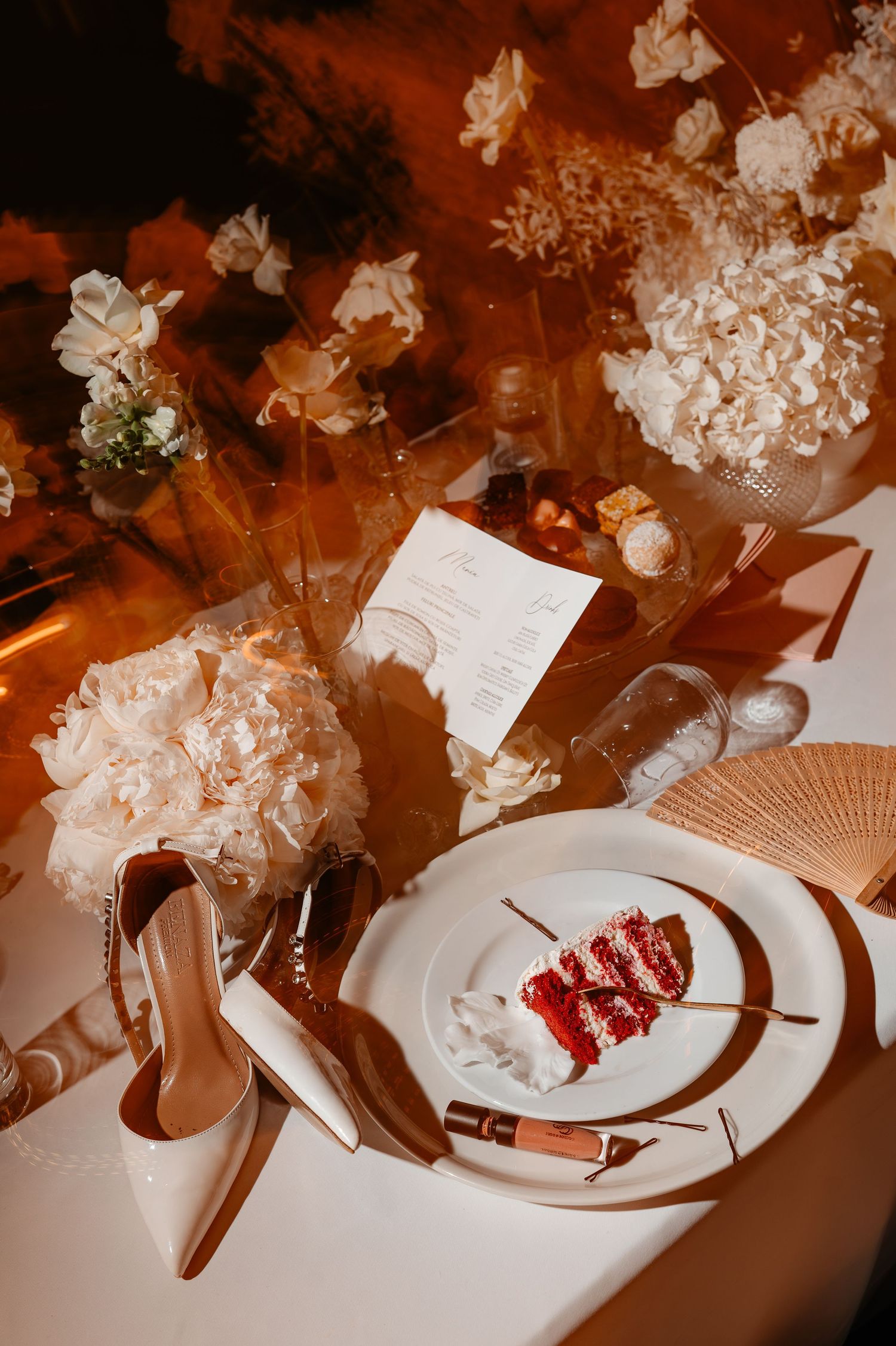 Wedding reception table detail with red velvet cake slice, white heels, peony bouquet, and gold accents.