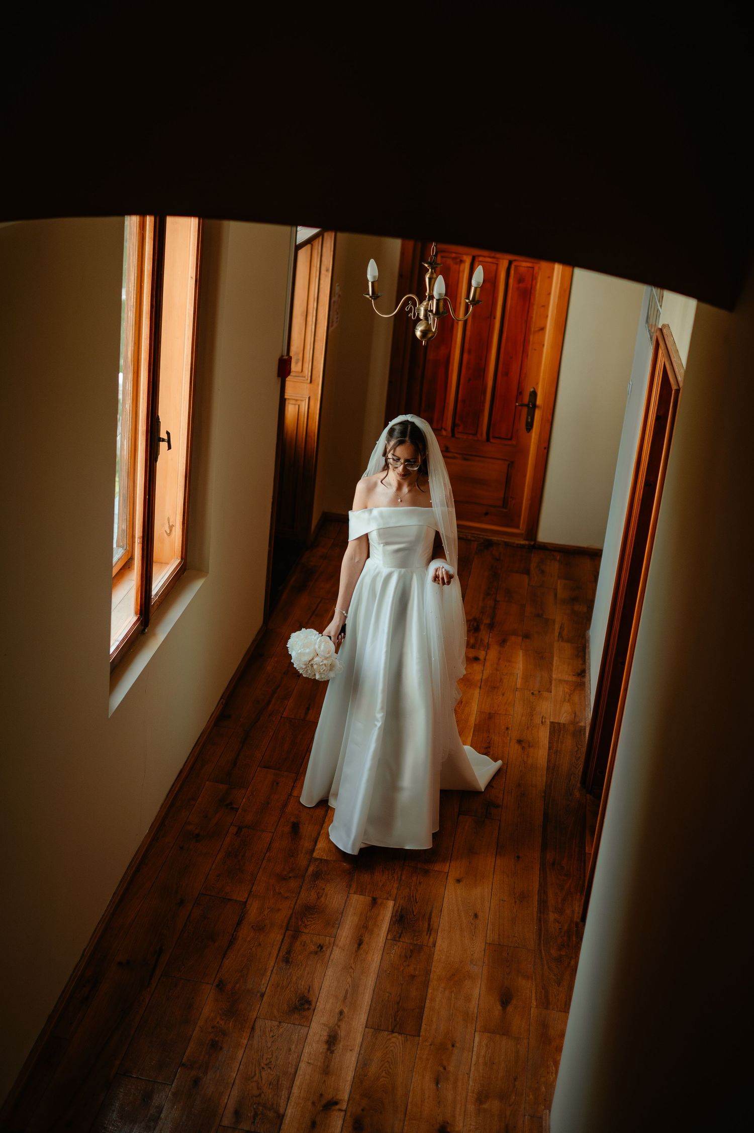 Aerial view of bride in white off-shoulder gown and veil walking through elegant hallway.