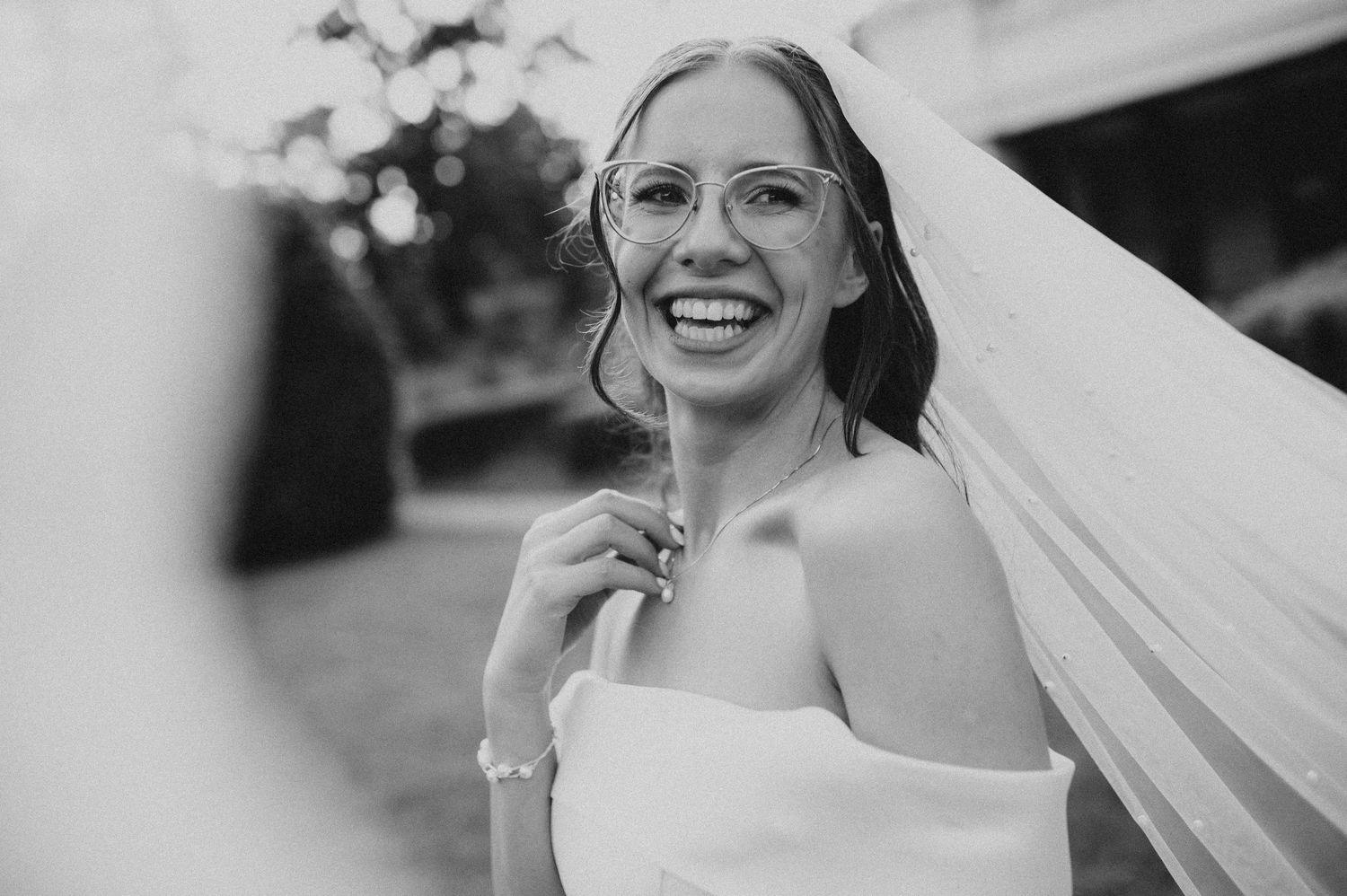 Smiling bride in glasses and veil laughing joyfully on her wedding day in a black and white photo.