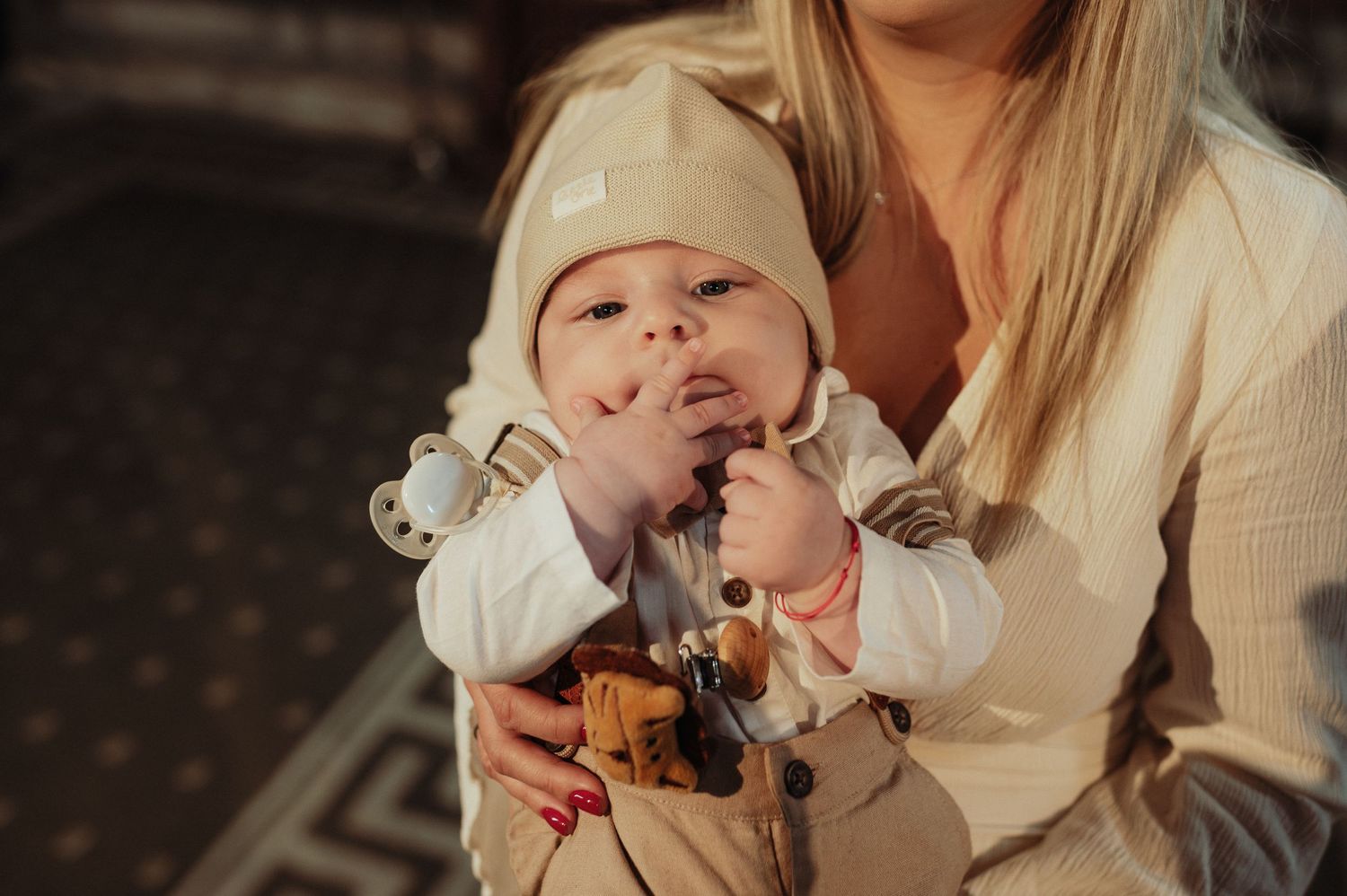 Close-up of a baby in a beige outfit and hat being held by mother inside a church.