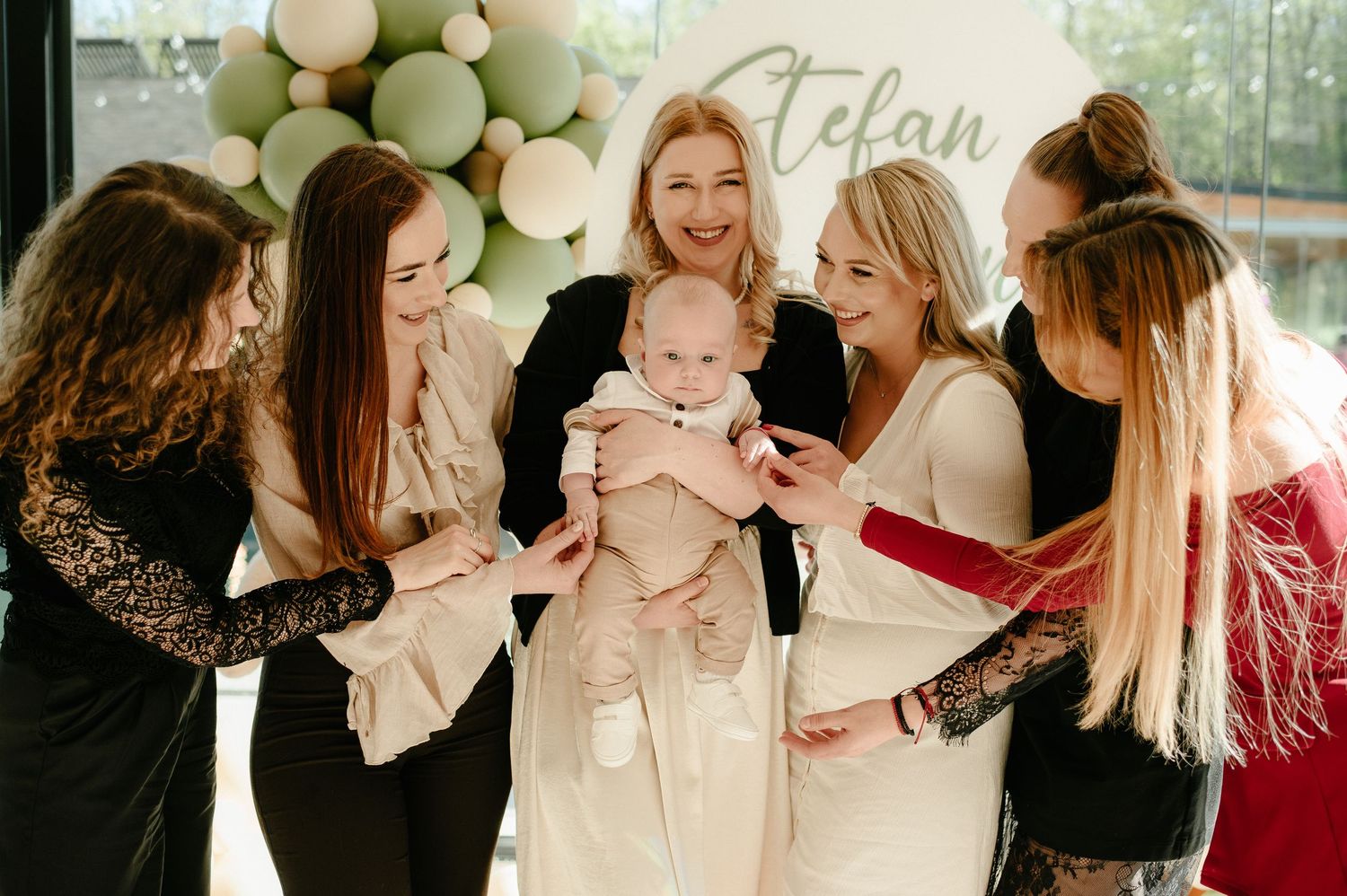 Group of smiling women gathered around a baby at a christening party with green balloon backdrop.
