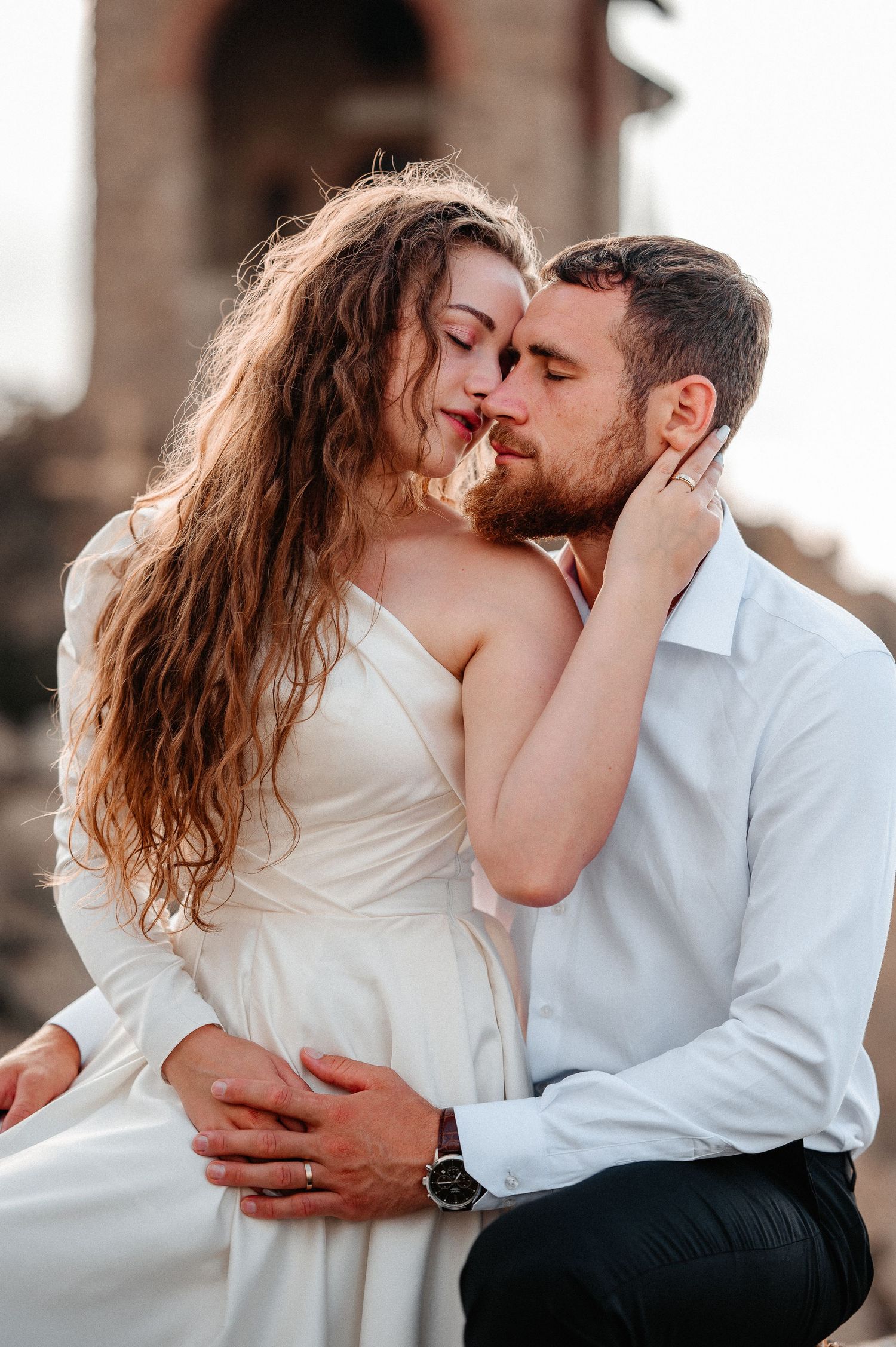 Bride in white dress and bearded groom sharing tender moment near ancient stone ruins.