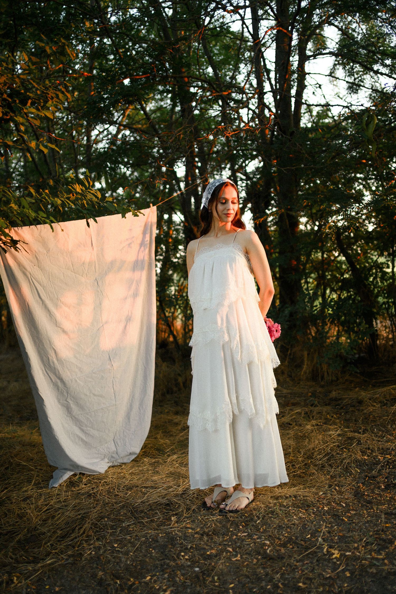 Elegant woman wearing white layered dress poses outdoors near draped cloth with trees in warm sunset light.