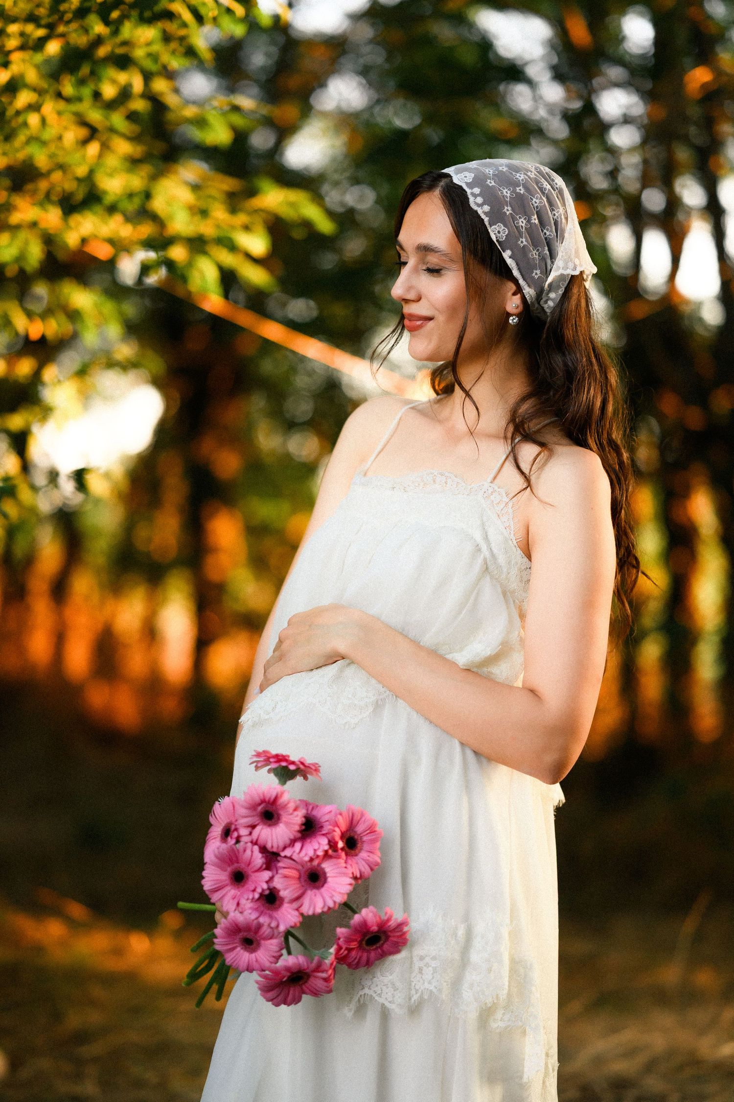 Pregnant woman in white dress holding pink gerbera daisies, wearing lace headscarf, standing in sunlit garden.