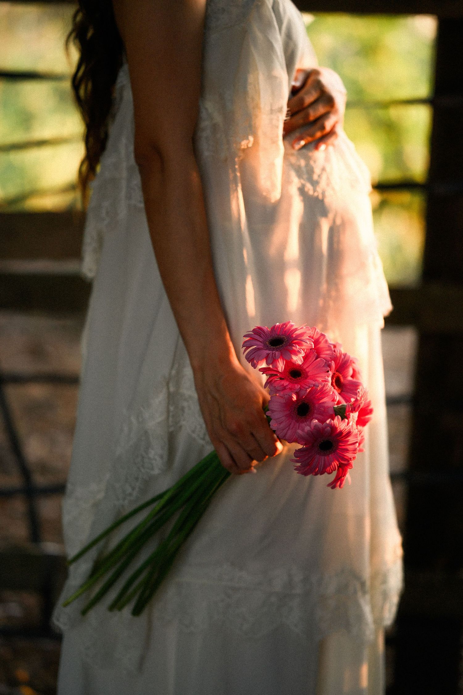 Bride in white lace dress holding pink gerbera daisy bouquet outdoors in warm sunlight.