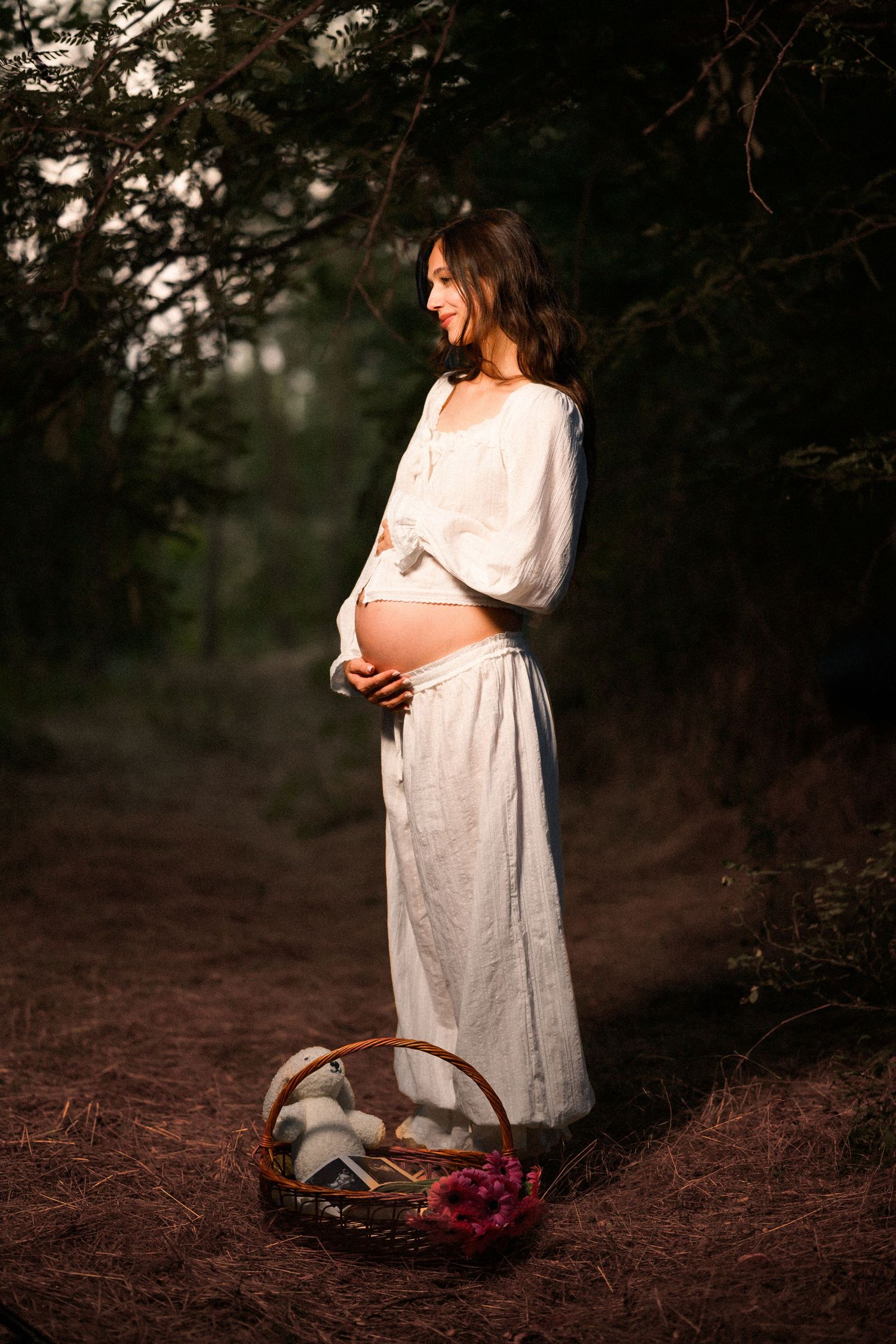 Pregnant woman in white dress holding her belly in a sunlit forest with a wicker basket of flowers at her feet.