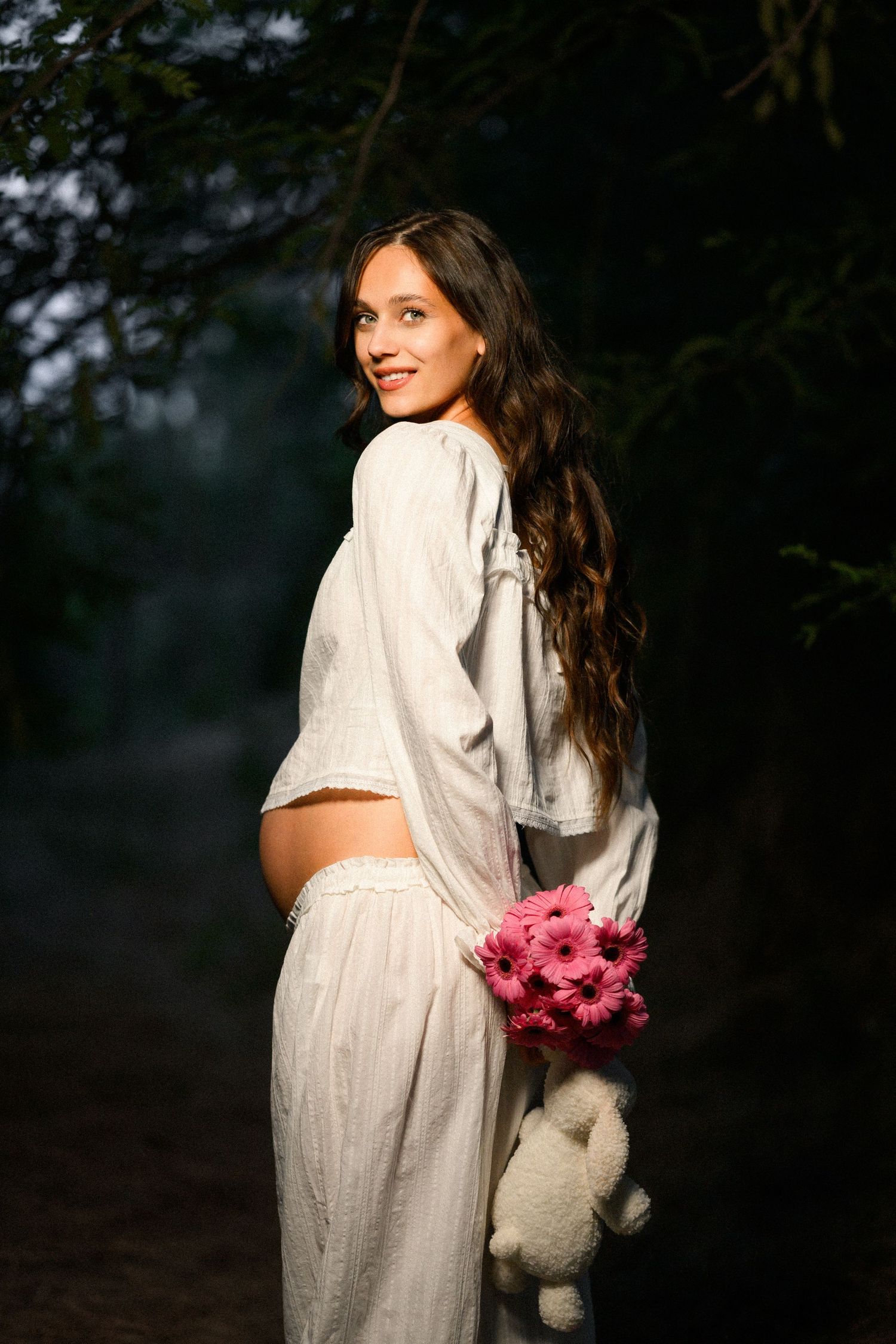 Pregnant woman in white outfit holding pink gerbera flowers and teddy bear, smiling in a dark forest setting.