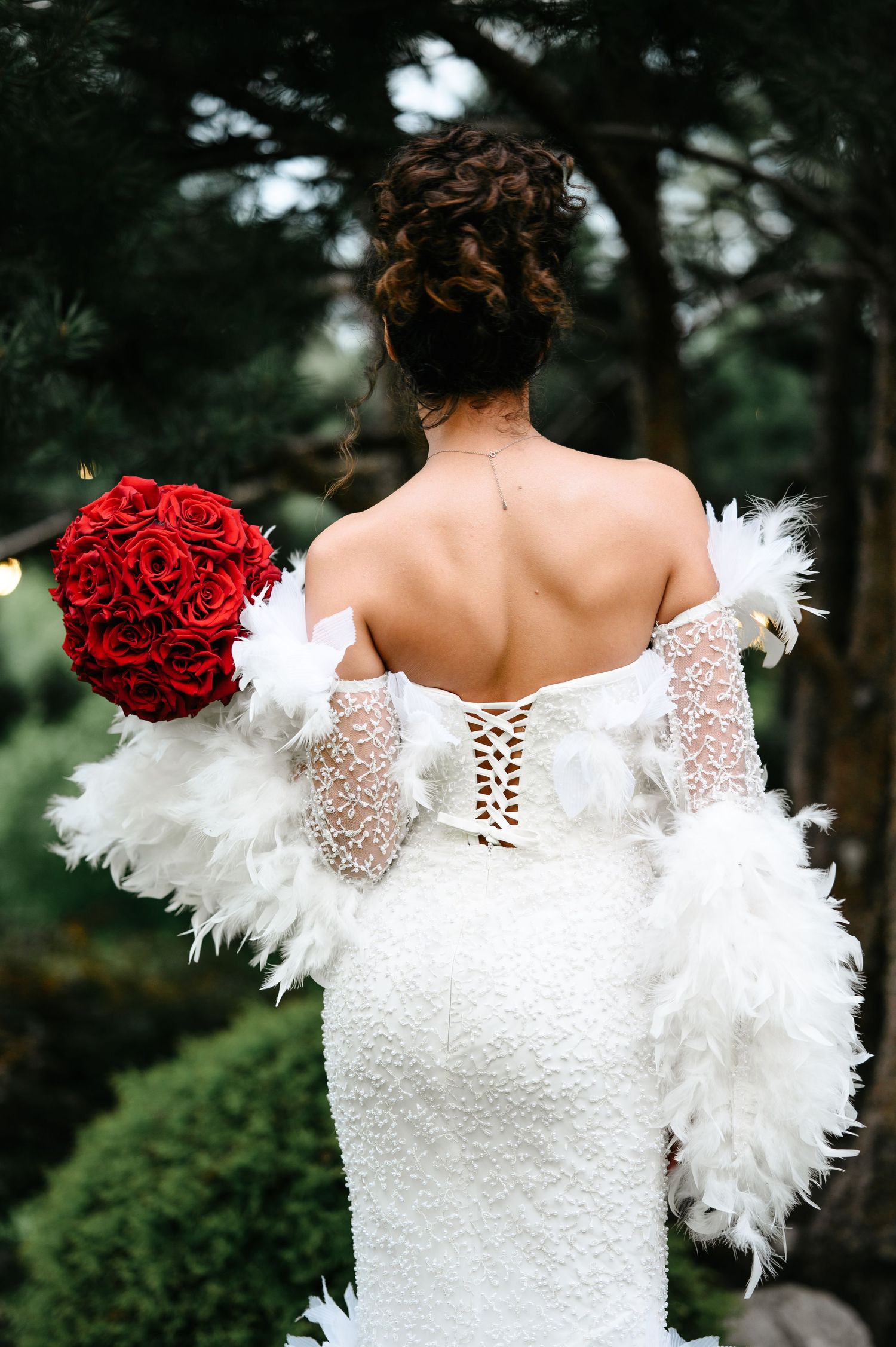 Bride in white feather wrap and lace corset gown holds red rose bouquet, viewed from behind outdoors.