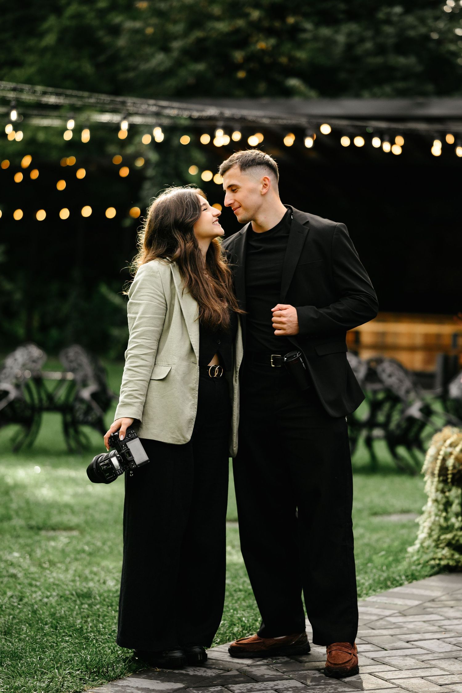 Romantic couple sharing a tender moment outdoors at night, woman holding camera, string lights glowing in background.