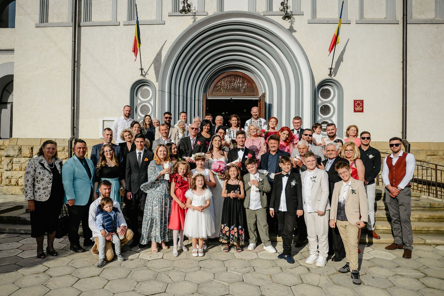 Large wedding group photo outside a white church with arched entrance, featuring guests in formal attire.