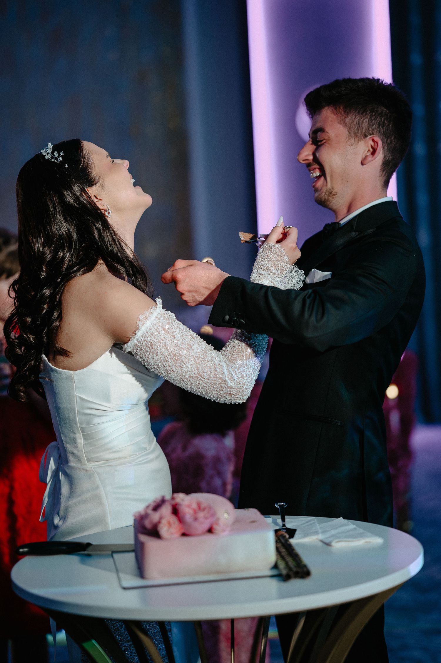Joyful bride and groom playfully feed each other wedding cake at their reception celebration.