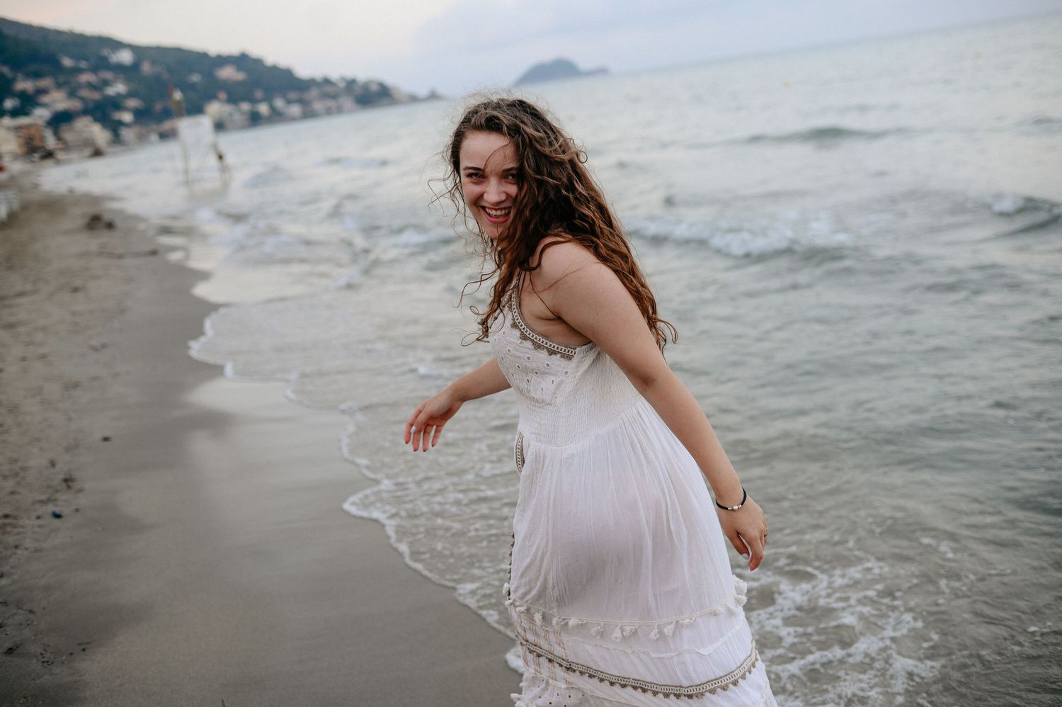 Joyful woman in white dress running along a sandy beach with ocean waves.