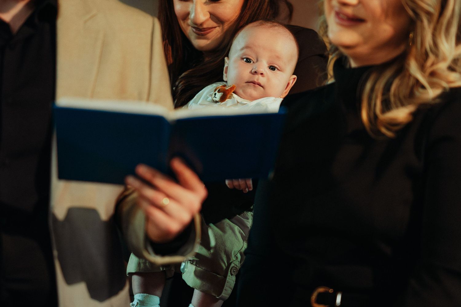A smiling woman holds a baby while another woman reads from a blue book at a baptism.