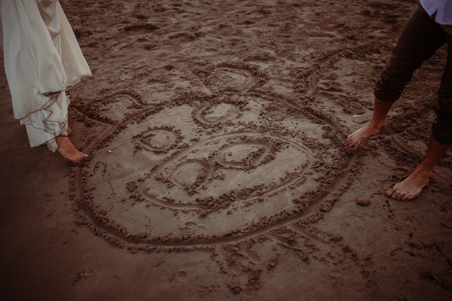 Couple standing barefoot on sand beside a heart and face drawing etched in the beach.