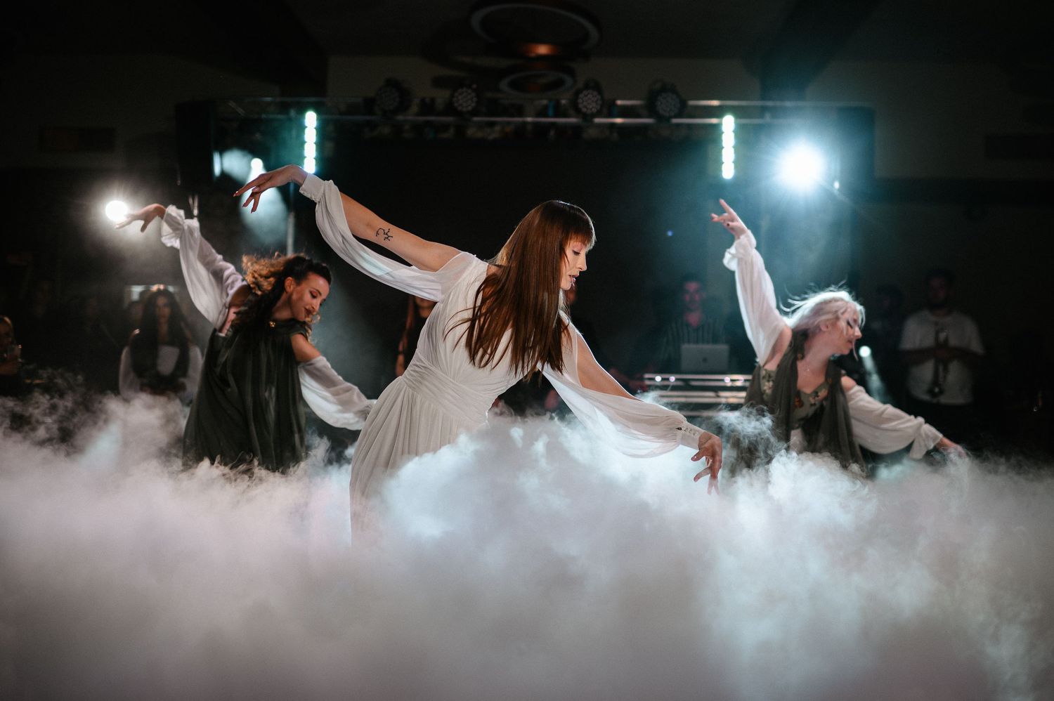 Female dancers performing elegant choreography in white dresses amid dramatic fog on stage.