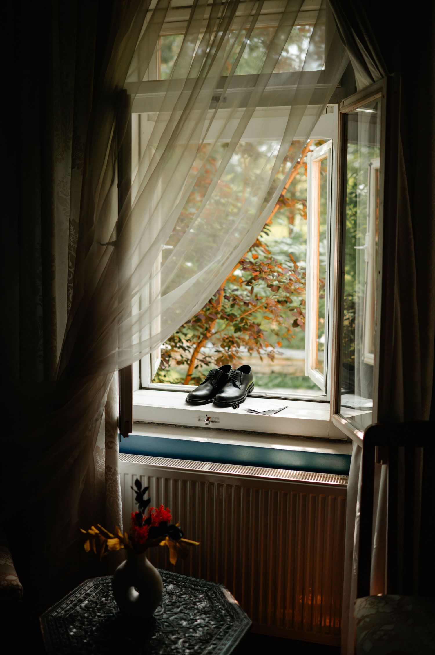 Black groom dress shoes displayed on sunlit window sill with sheer curtains and autumn foliage.