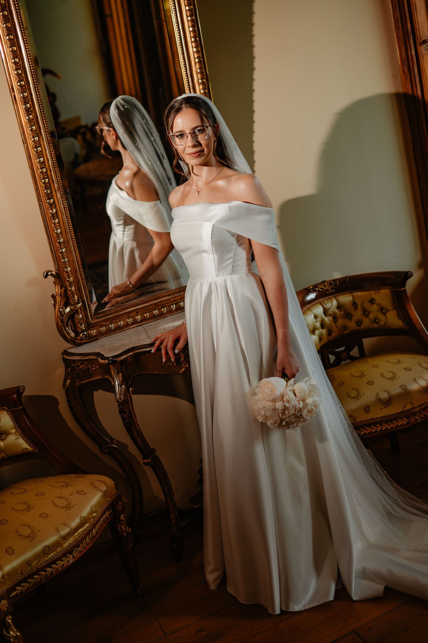 Bride in white off-shoulder satin wedding gown with veil and glasses stands before large gold-framed antique mirror.