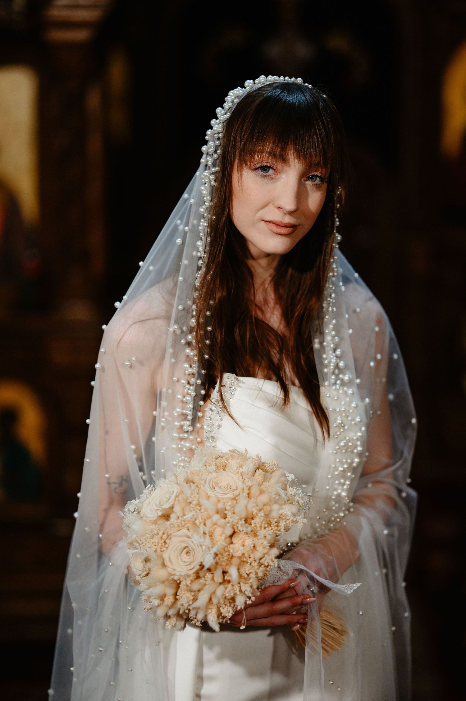 Elegant bride wearing pearl-adorned veil holds cream rose bouquet inside ornate Orthodox church.