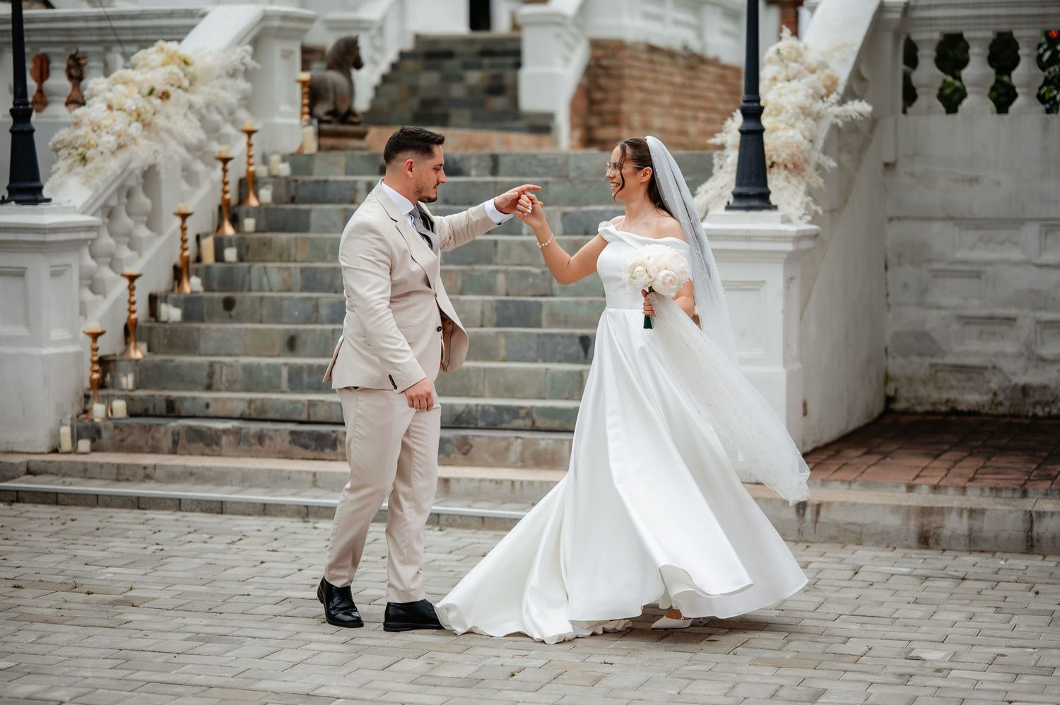Groom twirls bride in white wedding gown on cobblestone courtyard outside white neoclassical wedding venue with staircase.