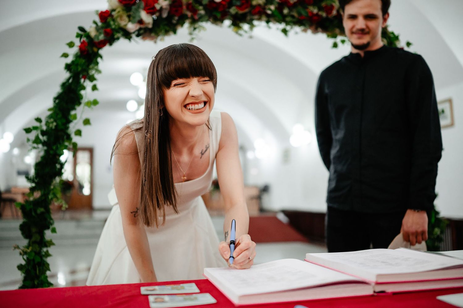 Smiling bride in white dress signs wedding registry at red-draped table beneath floral arch of red roses.