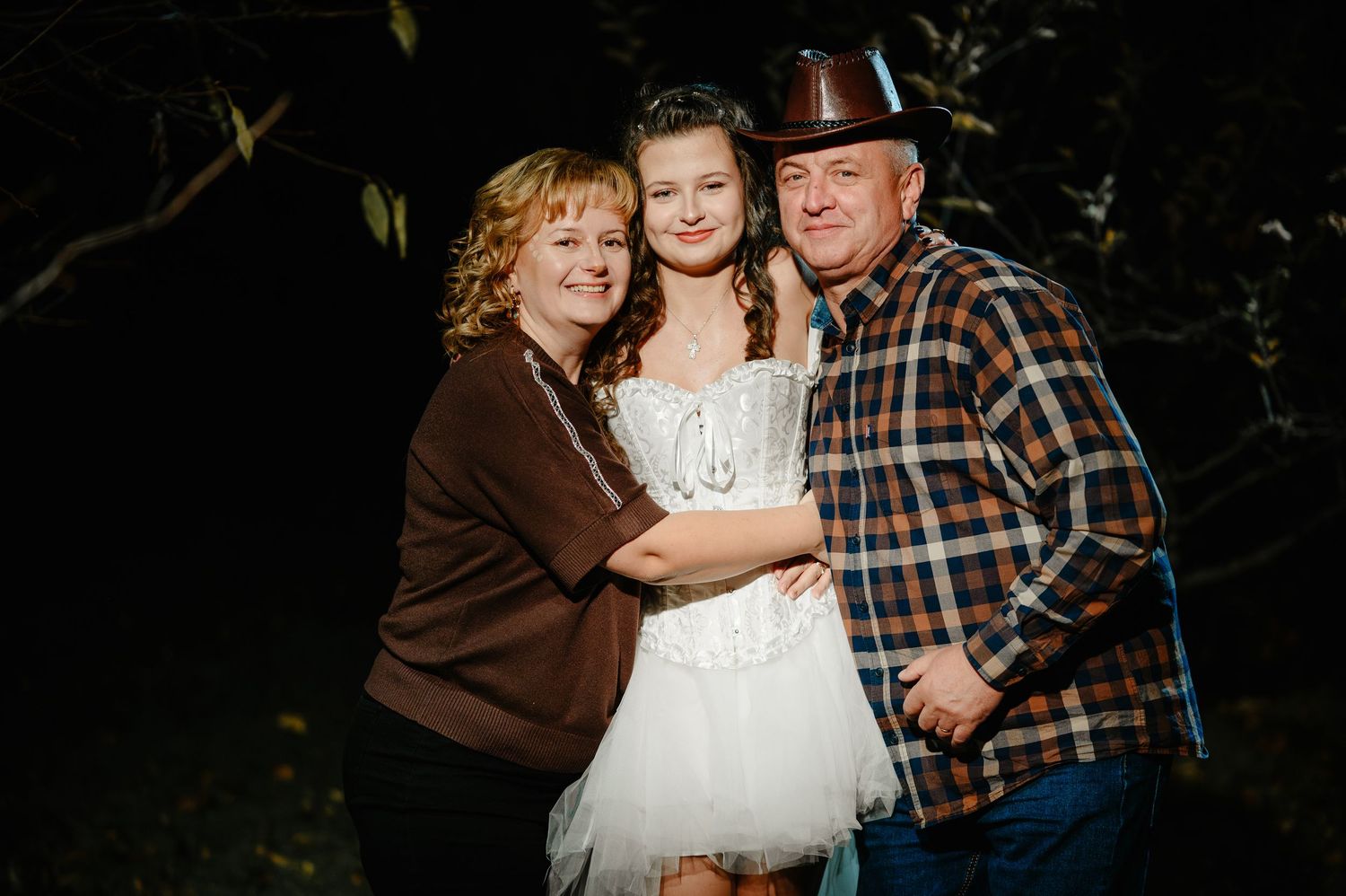 Family of three posing outdoors at night, woman in brown top, girl in white dress, man in cowboy hat.