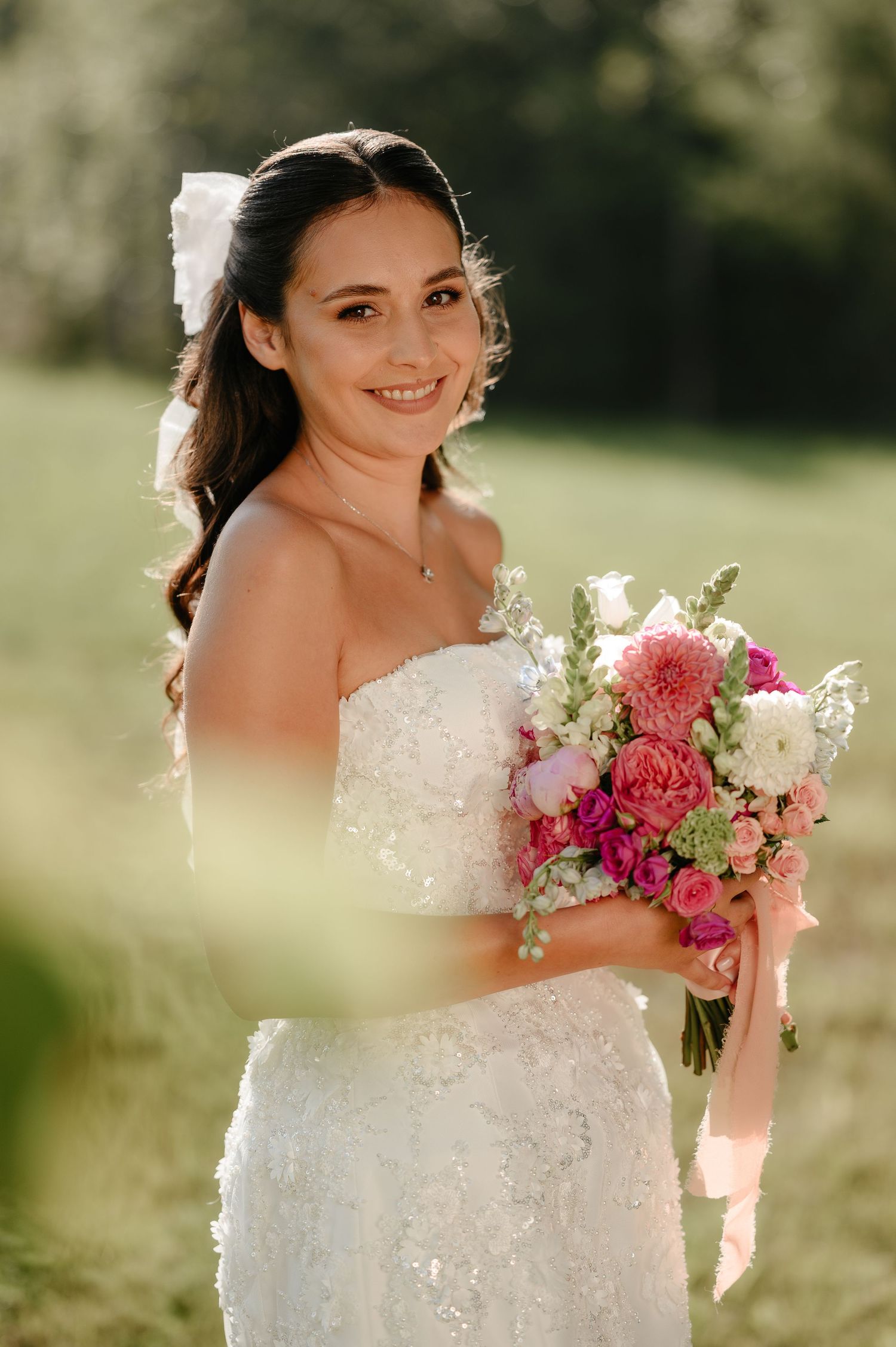 Smiling bride in white lace dress holding a pink and white bouquet in a sunlit garden.