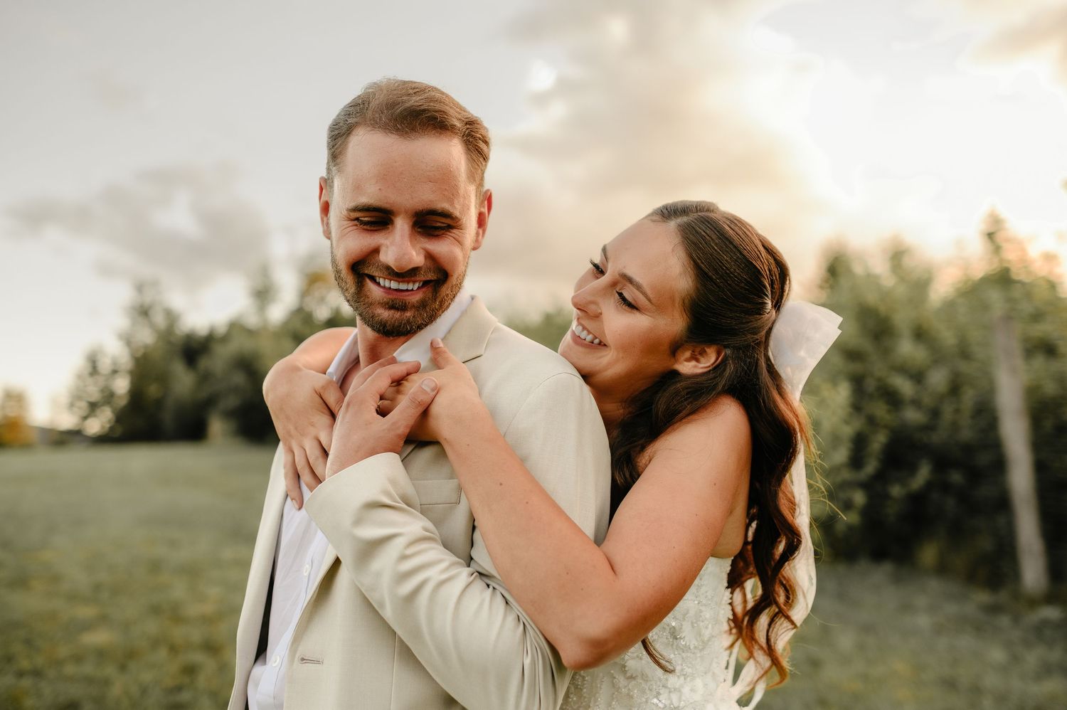 Smiling groom in beige suit embraced from behind by laughing bride in sunny outdoor setting.