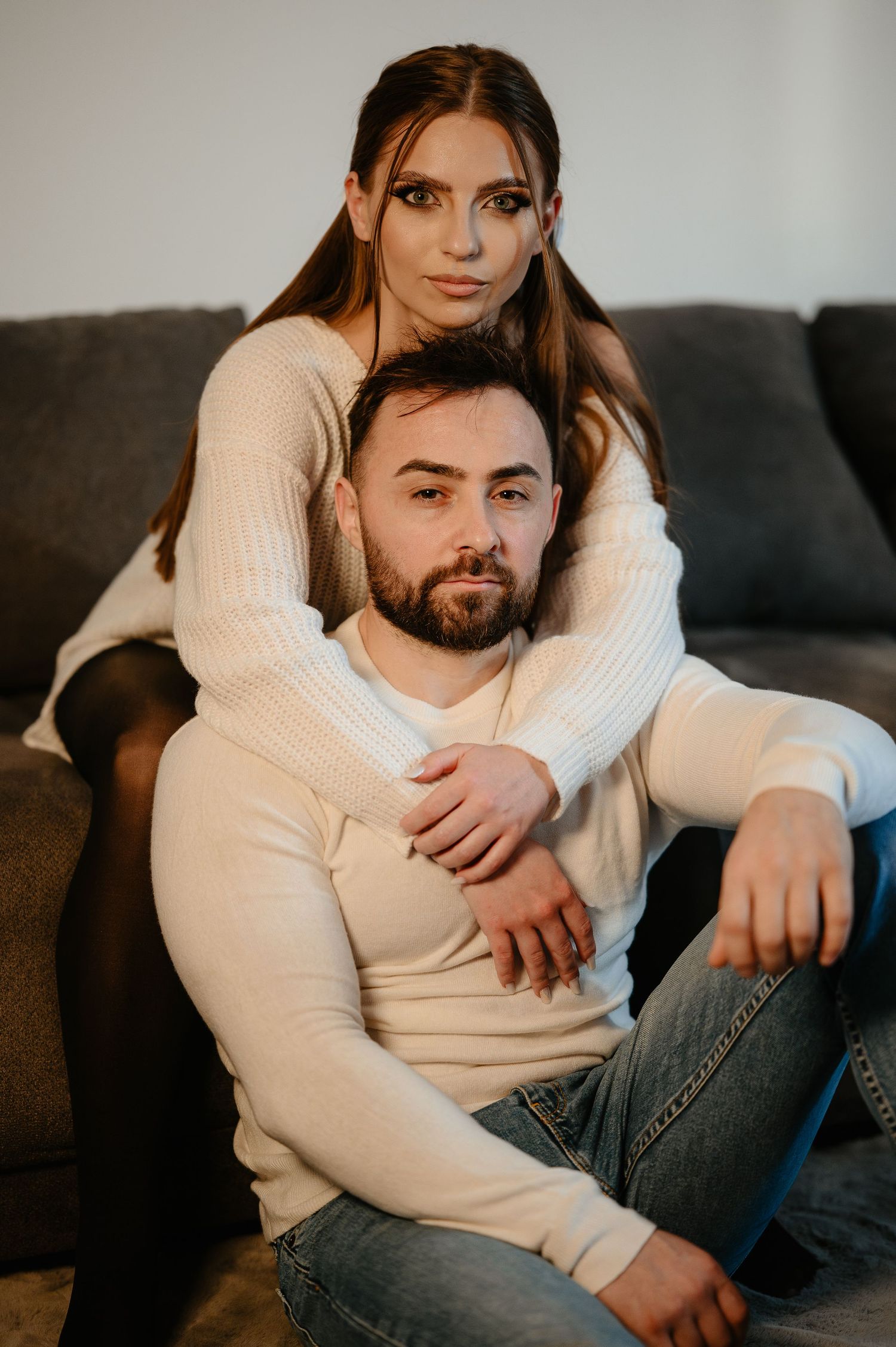 Young couple posing together by a gray sofa, woman embracing man from behind affectionately.