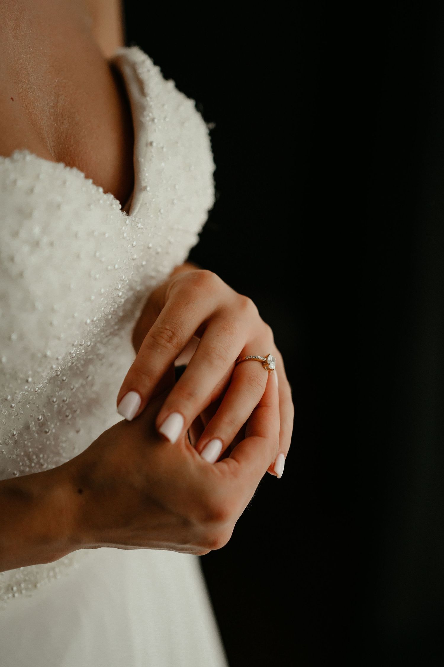 Close-up of bride's hands with white manicure and diamond engagement ring against beaded wedding dress.