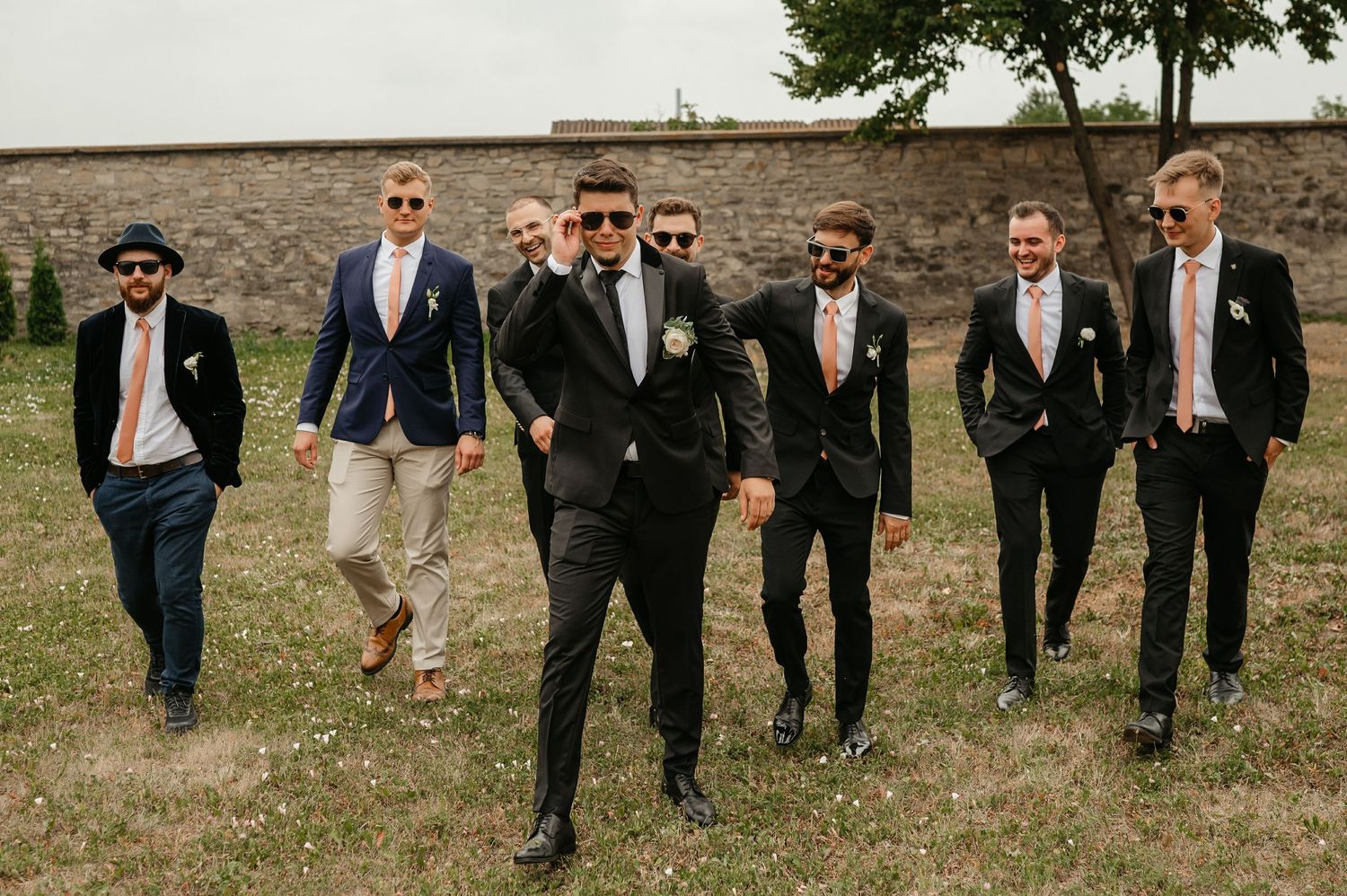 Groomsmen walking confidently in suits with pink ties toward the camera at an outdoor wedding venue.